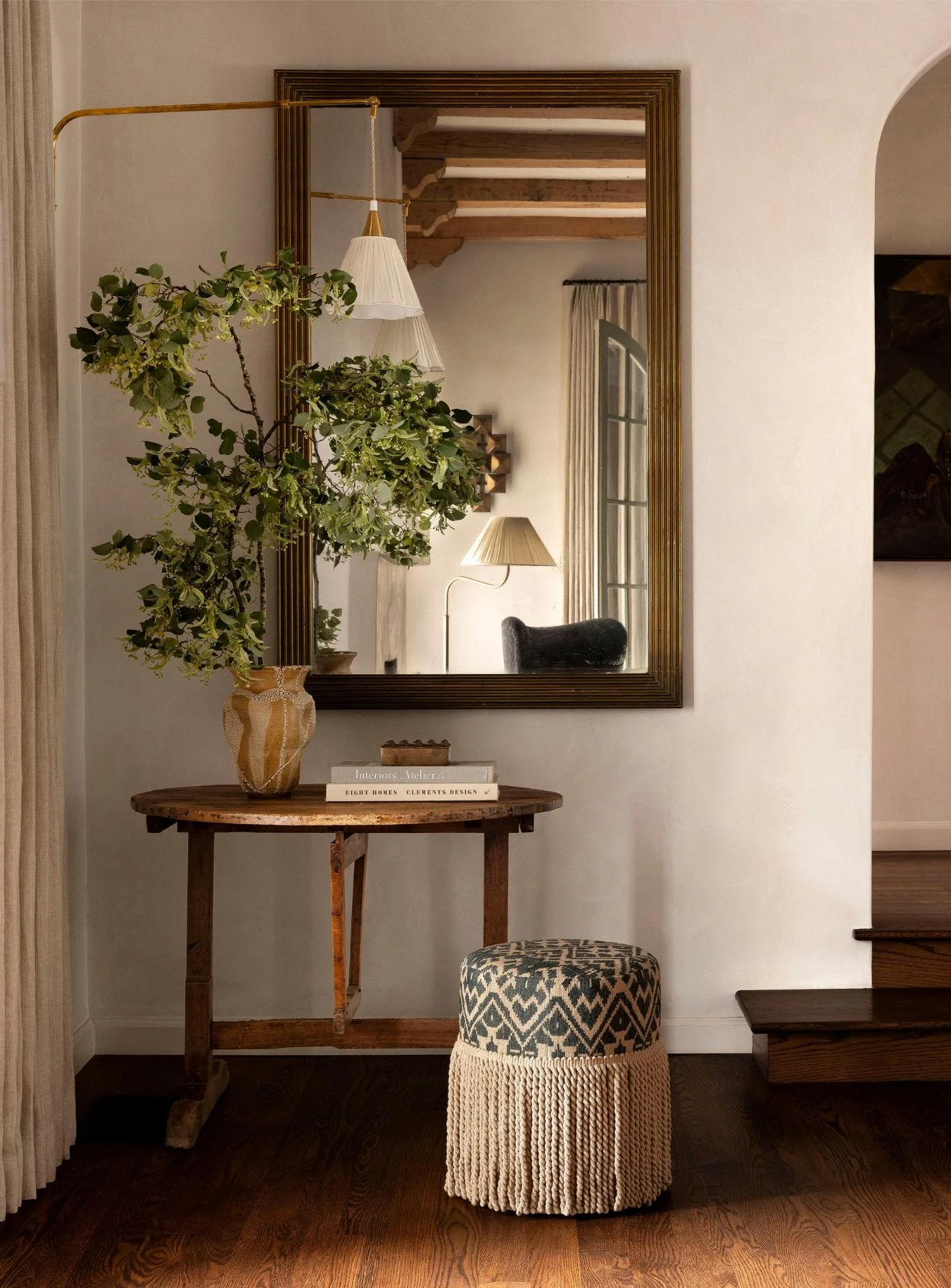 A rustic round wooden table holds a leafy vase below a large bronze-framed mirror, flanked by a fringed geometric ottoman in a warm hallway.