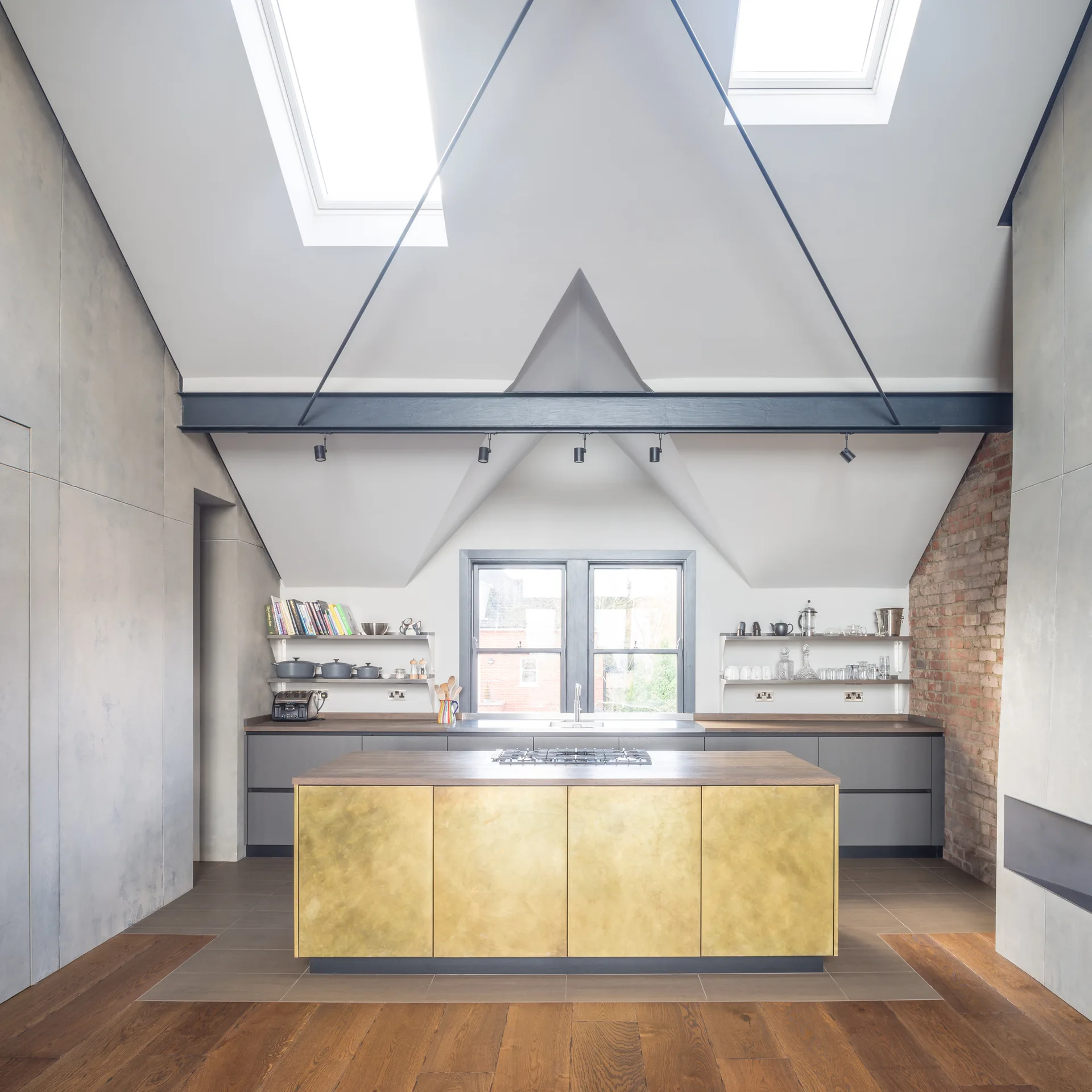 A modern loft kitchen featuring a central gold brass island, sloped ceilings with skylights, grey cabinetry, and a black structural beam.
