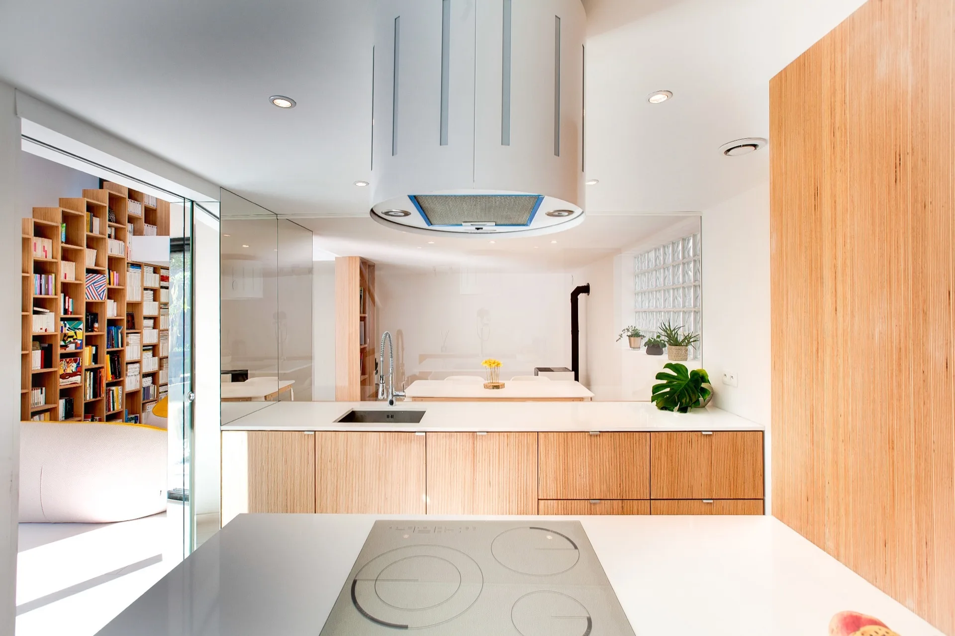 A modern kitchen featuring light plywood cabinetry, a white countertop, an integrated sink, and a large cylindrical range hood hanging from the ceiling.