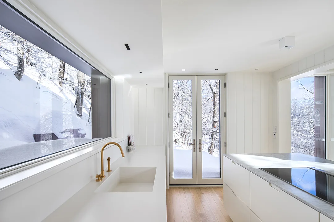 A minimalist white kitchen featuring an integrated sink with a gold faucet, flat-panel cabinets, and a large window looking out at a snowy forest.