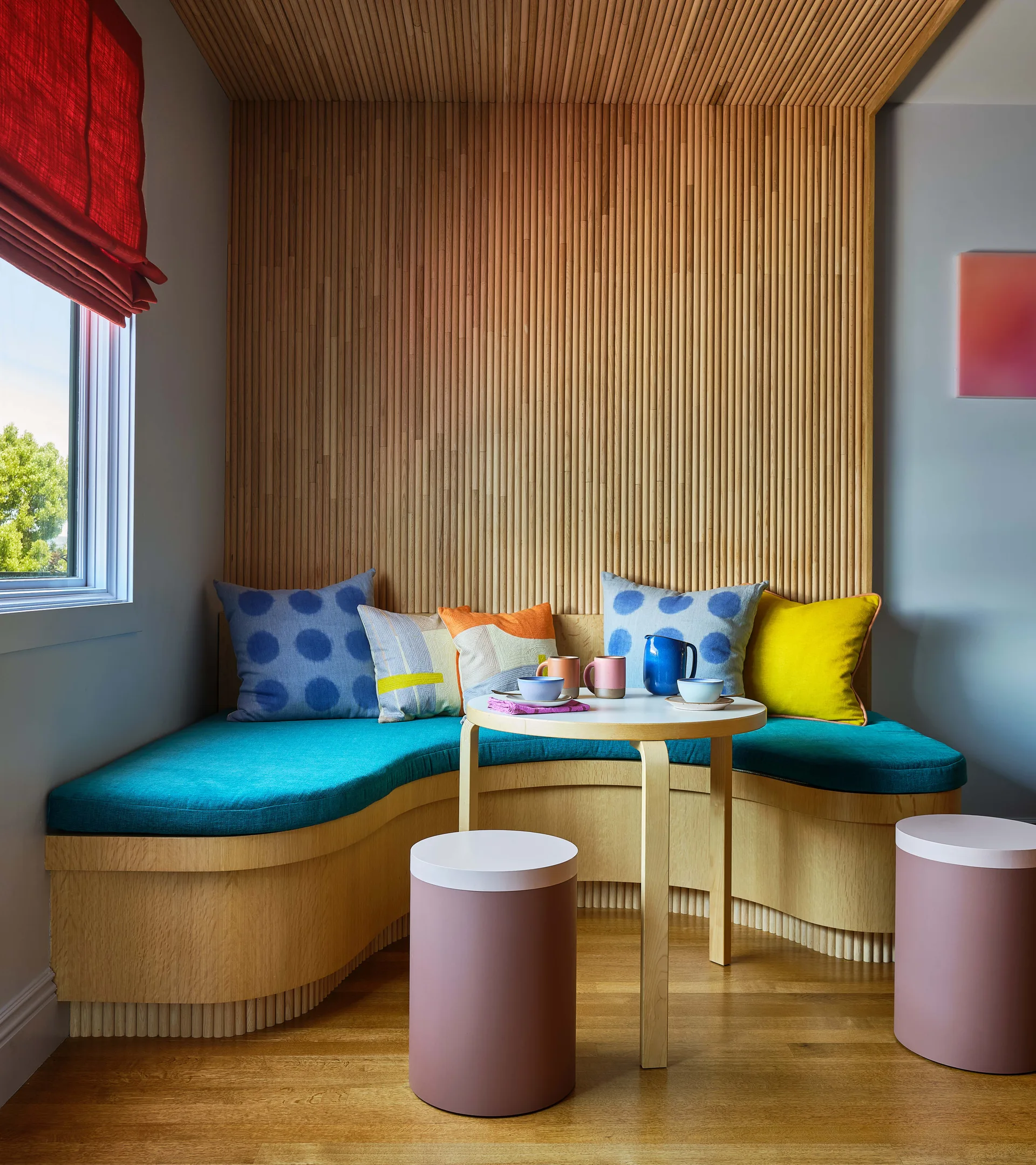 A dining nook with a curved wood banquette, teal cushions, vertical slat paneling, a round wood table, and two pink cylinder stools.