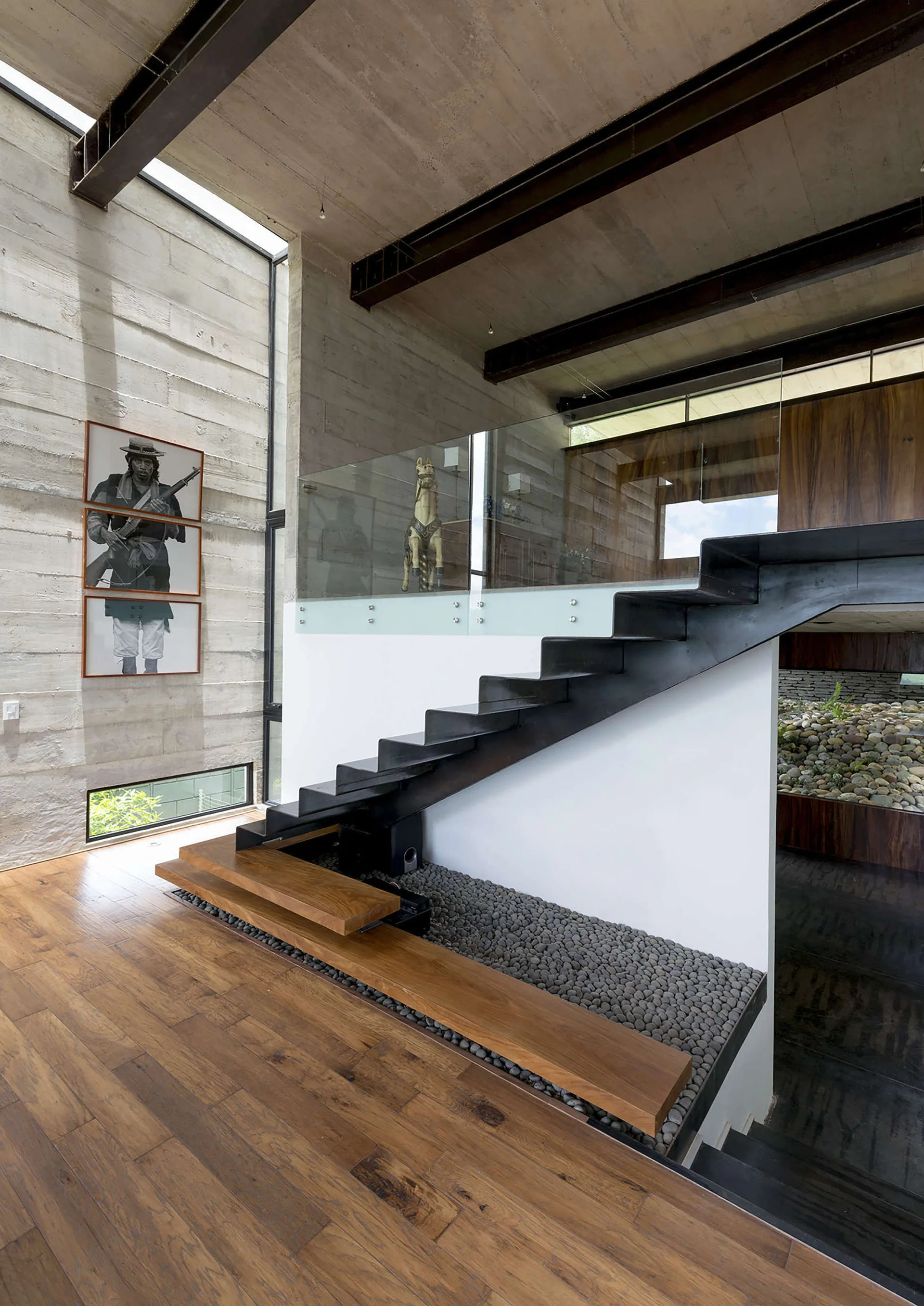 A modern black metal floating staircase rises next to a textured board-formed concrete wall and a decorative grey pebble bed inlay under the steps.