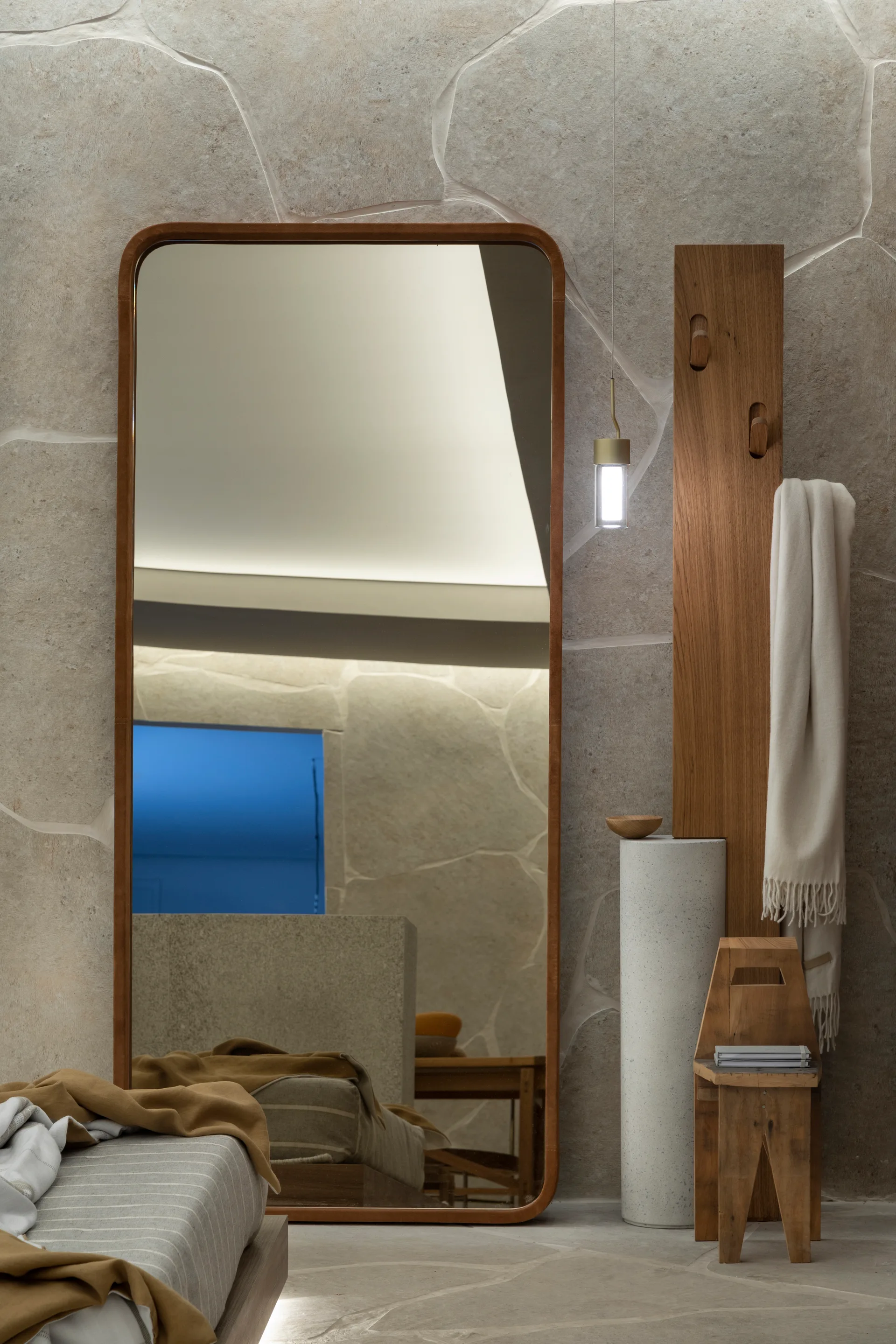 A tall wood-framed floor mirror stands against a textured beige stone wall next to a wooden wall-mounted coat rack and a white speckled pedestal.