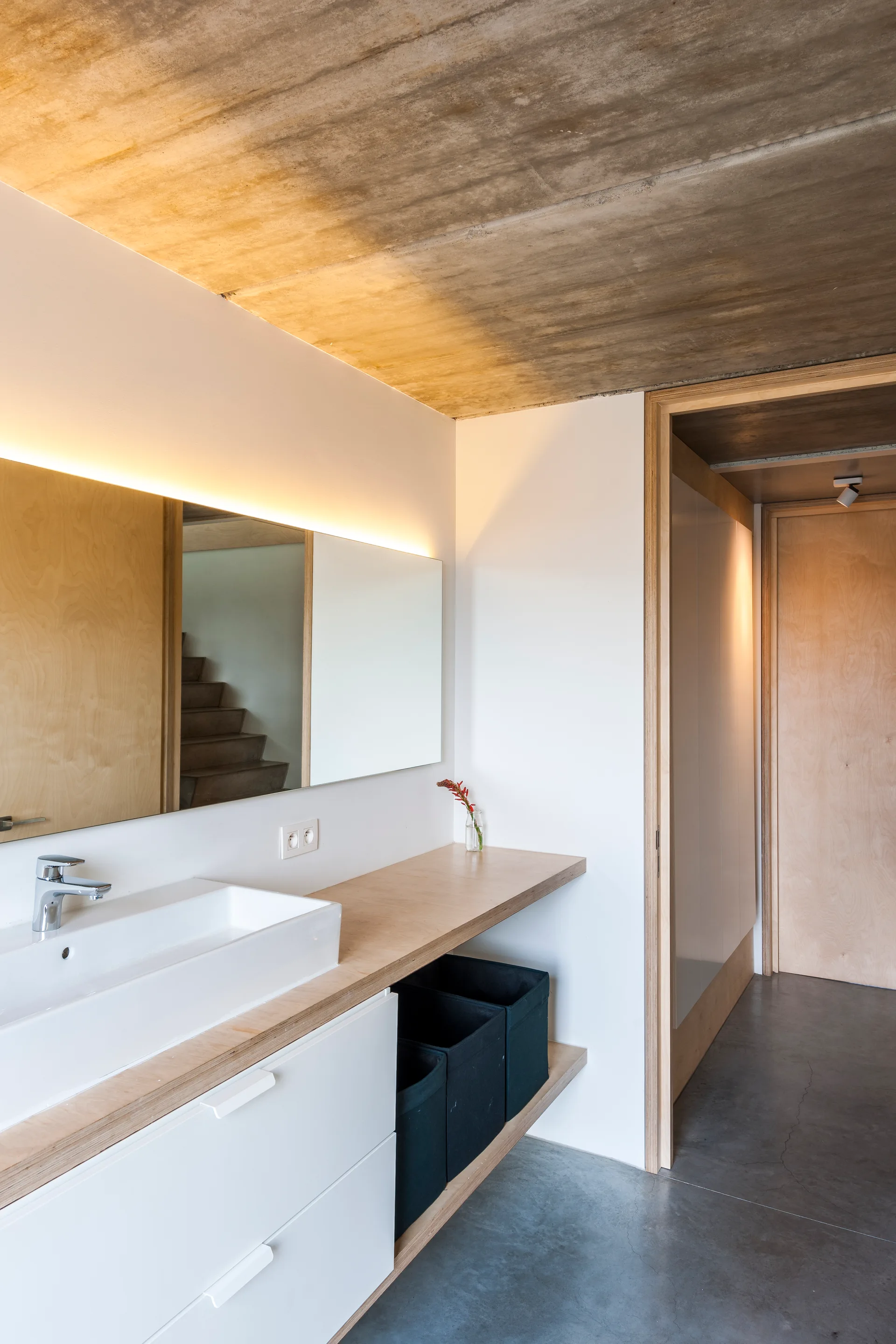 A minimalist bathroom features a white vanity with a plywood countertop, a vessel sink, a long back-lit mirror, and an exposed raw concrete ceiling.