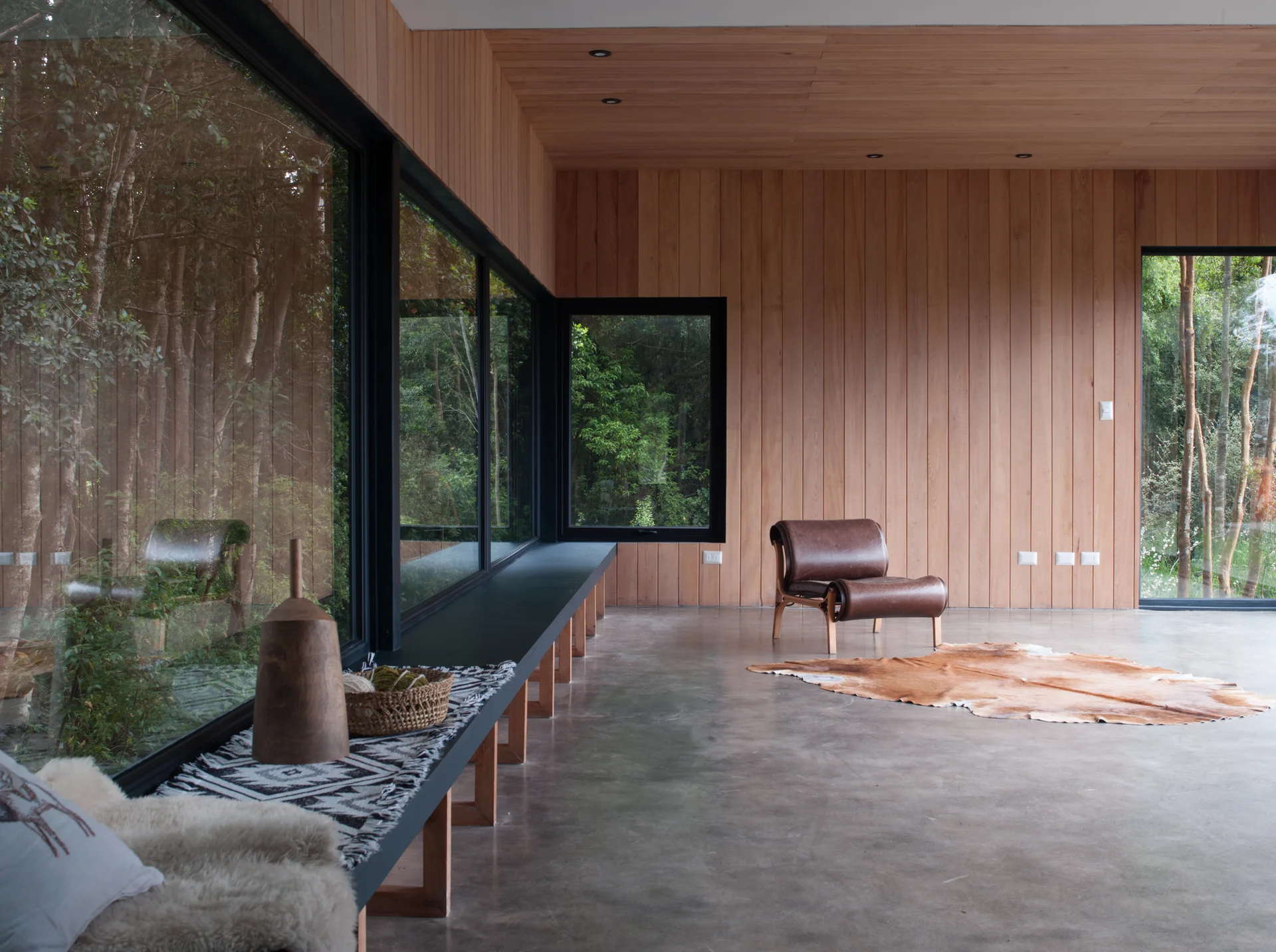 A minimalist room with vertical wood paneling, polished concrete floors, large windows showing a forest, and a single brown leather armchair on a cowhide rug.