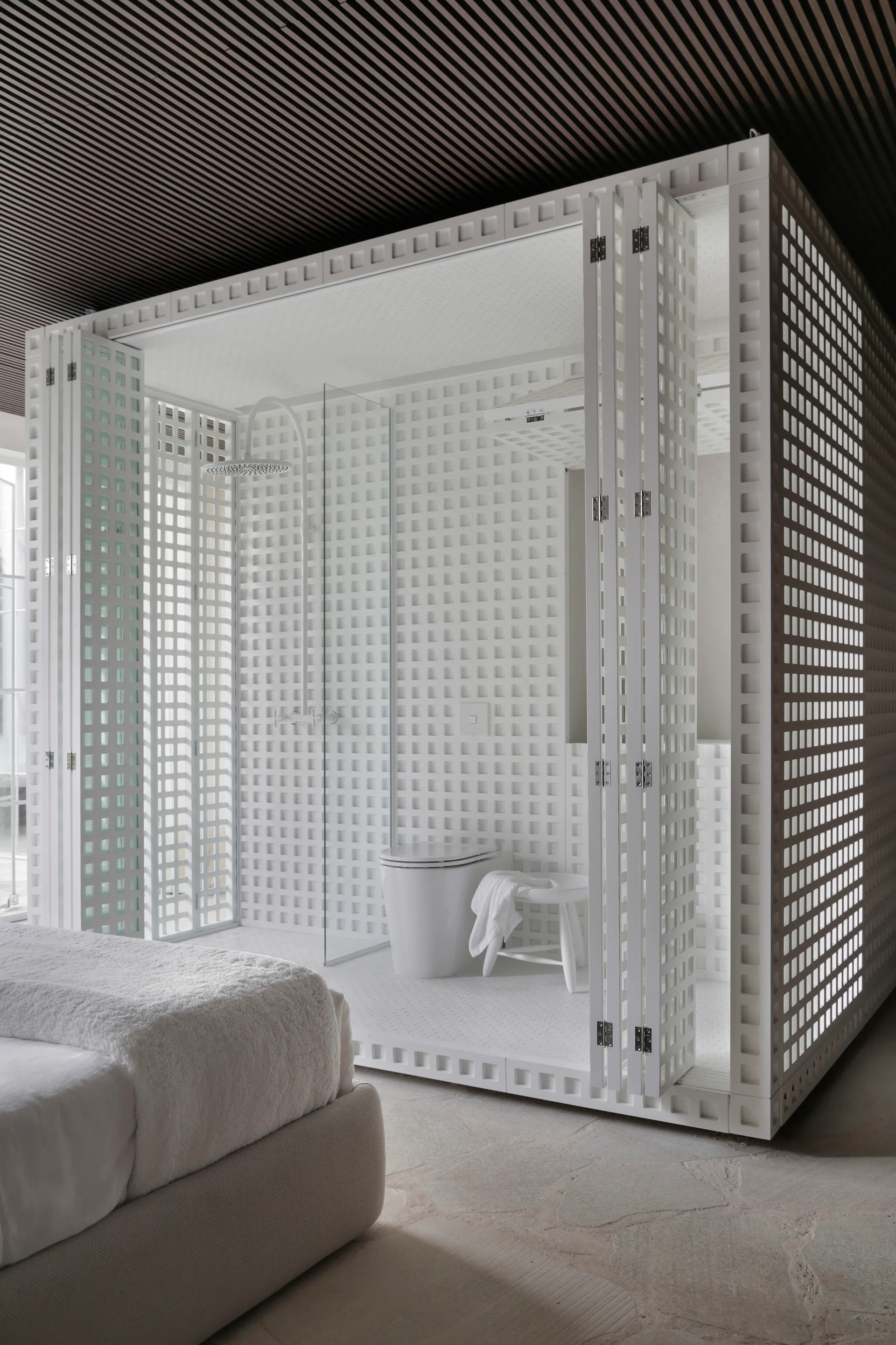 A white grid modular bathroom enclosure sits adjacent to a bed on flagstone flooring beneath a dark slatted ceiling.