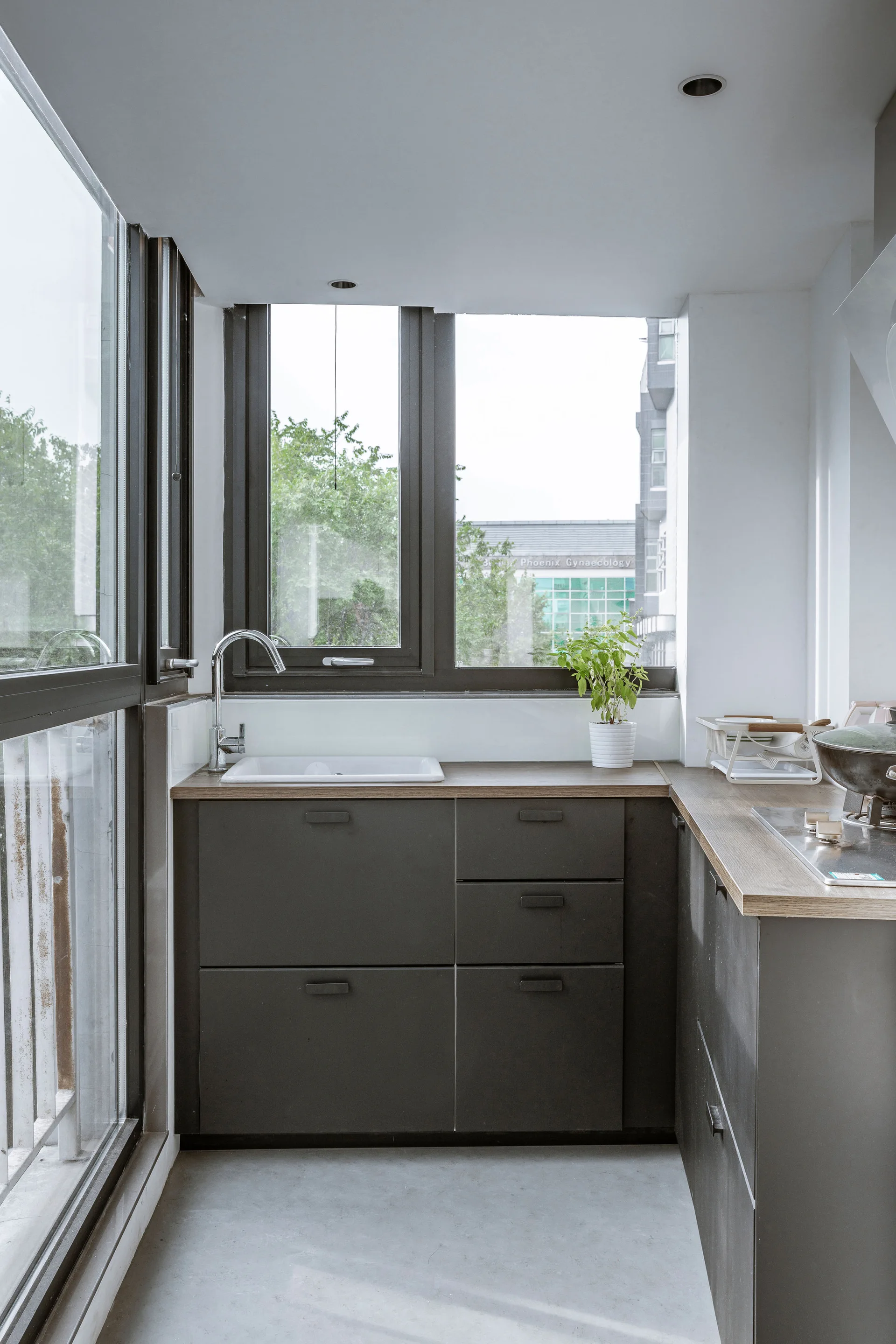 A minimalist kitchen featuring dark grey cabinets with a wood-toned countertop, a white ceramic sink beneath large windows, and a concrete-look floor.