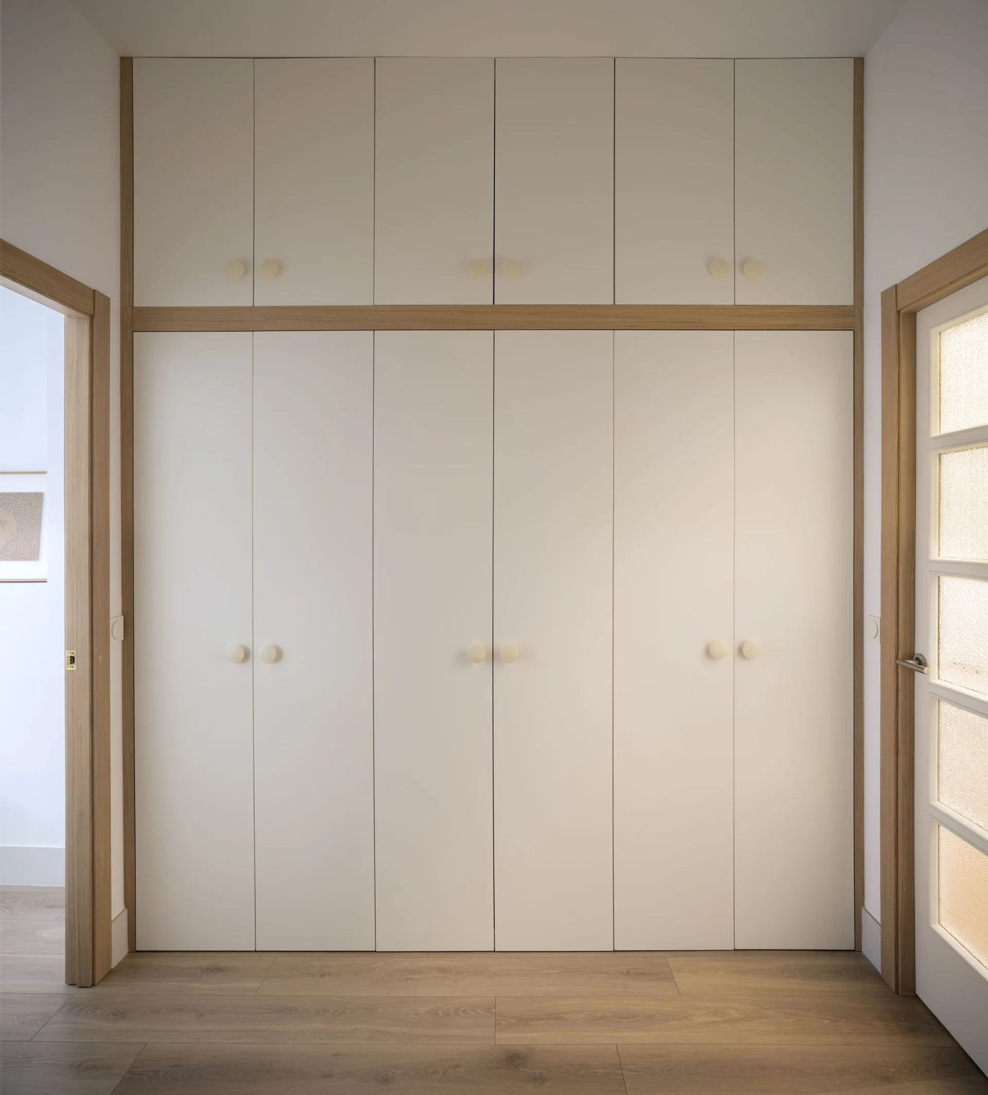 A floor-to-ceiling built-in wardrobe with off-white doors, round wooden knobs, and a light wood trim, set between two doorways on a wooden floor.