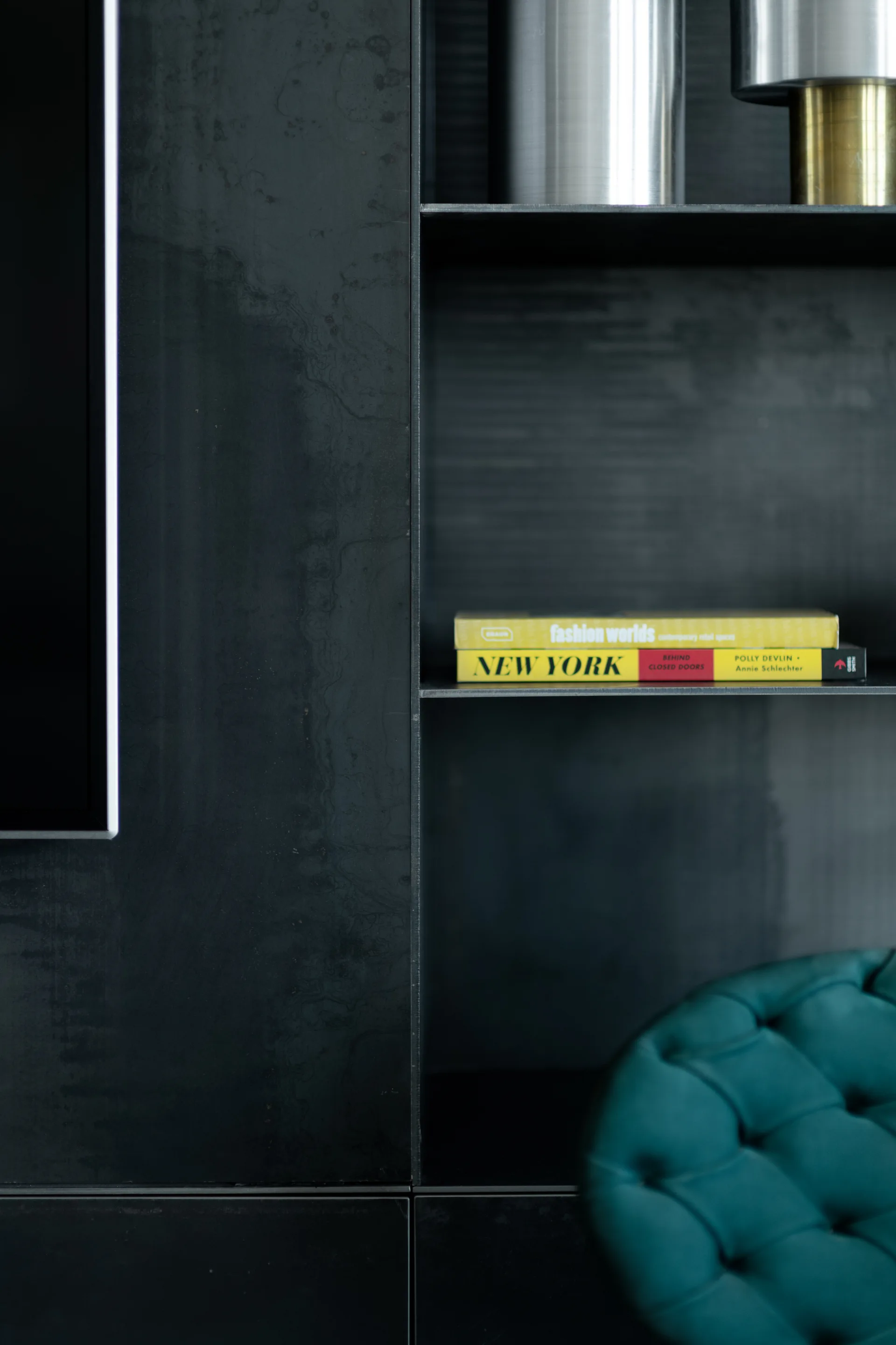 A dark charcoal metal shelving unit with yellow books sits next to an emerald green tufted velvet chair.