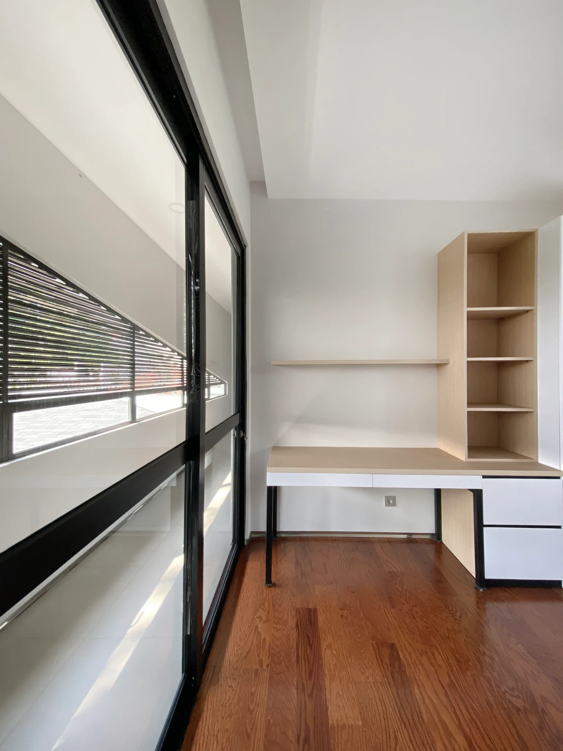 A minimalist home office with a light wood and white built-in desk, built-in shelving, warm hardwood floors, and a large window with black frames.