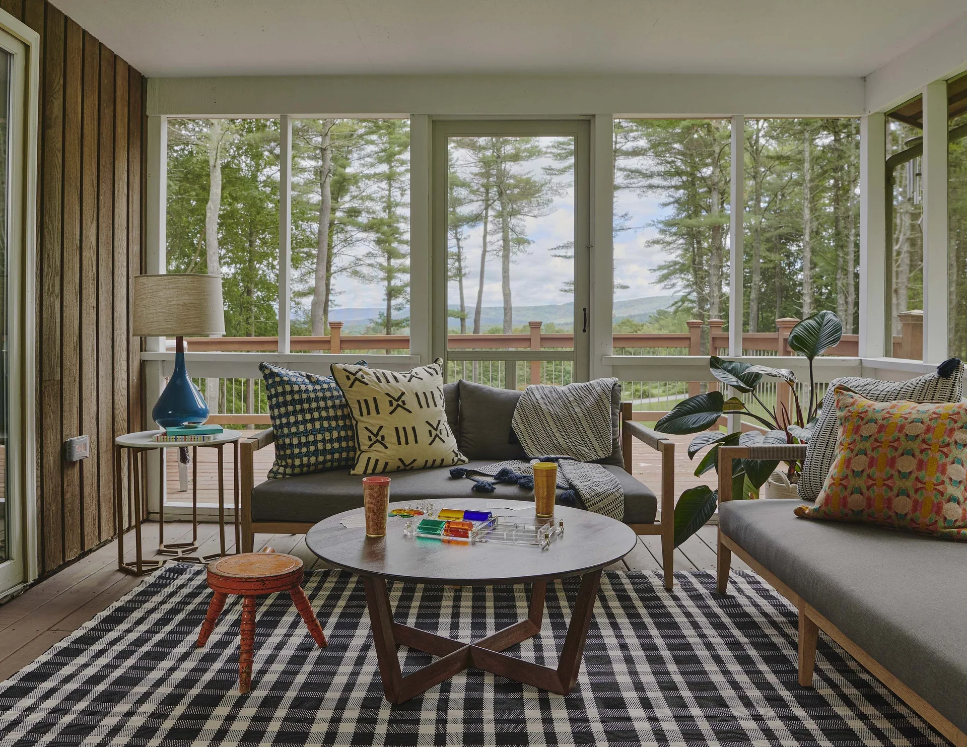 A screened-in porch with two gray sofas and a round wooden coffee table set on a black and white checkered rug with trees visible outside.
