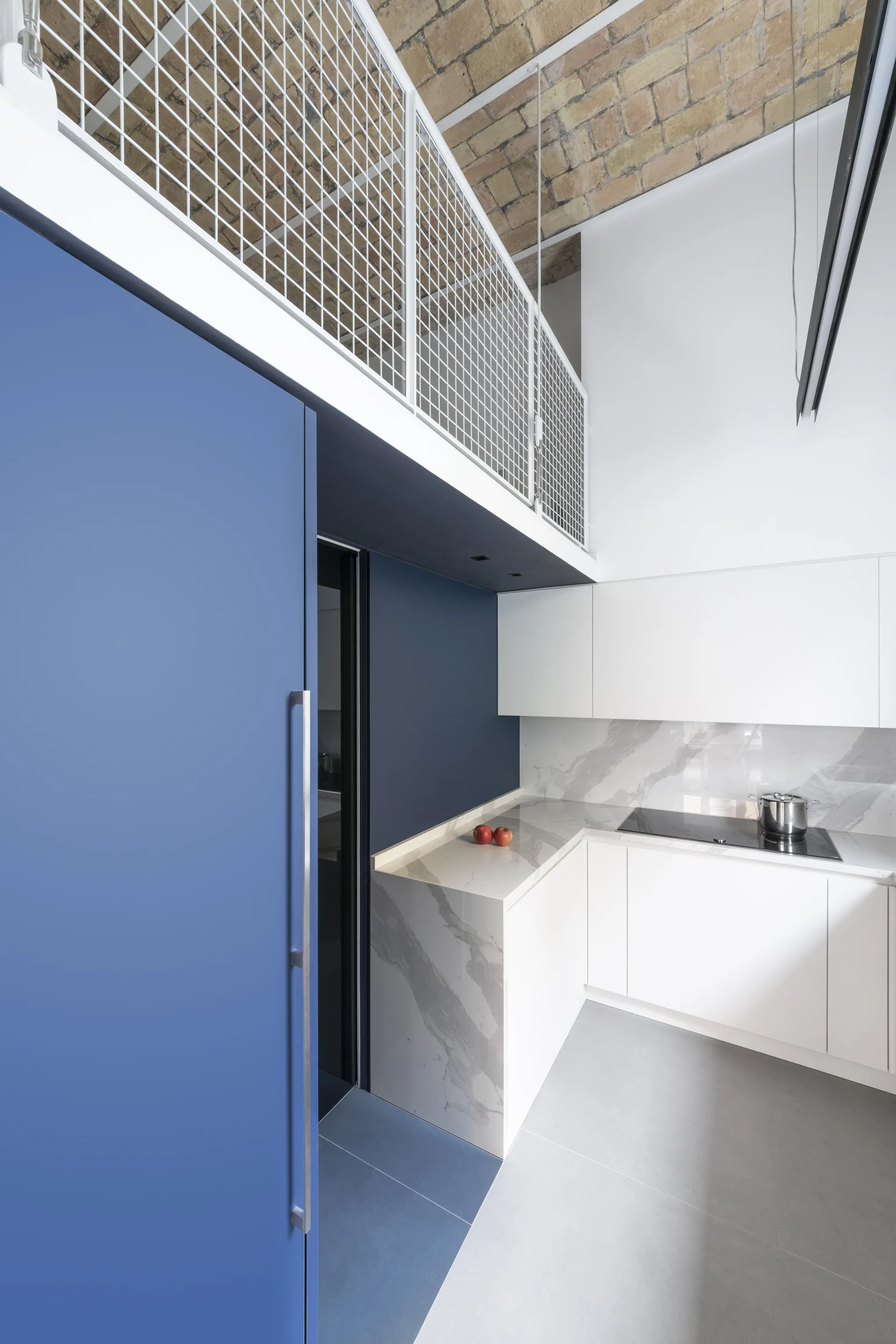 A modern kitchen with minimalist white cabinets, marble countertops, a bold blue wall panel, and an exposed brick vaulted ceiling below a mezzanine loft.