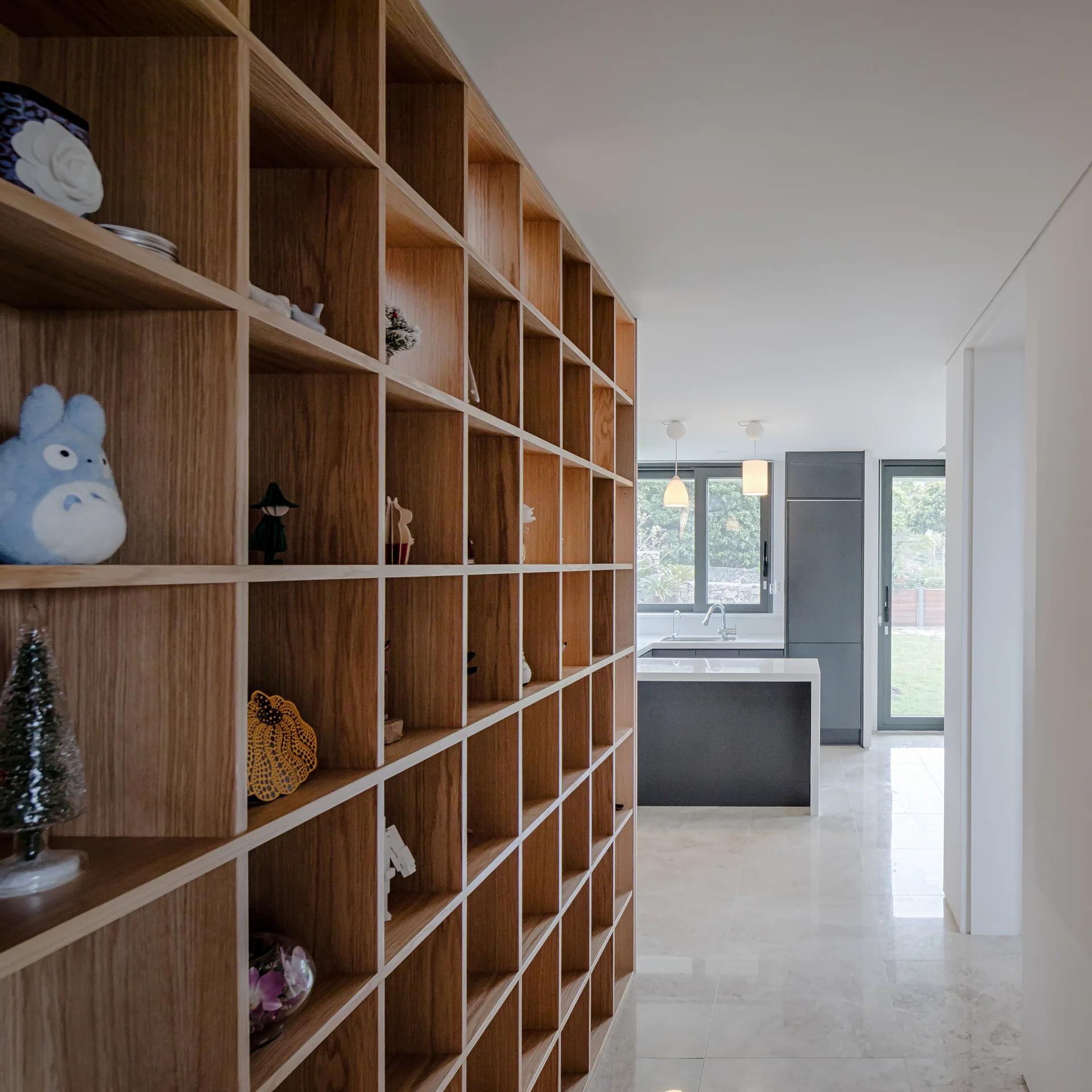 A large light wood grid shelving unit acts as a partition between a glossy tile hallway and a modern kitchen with grey cabinets.