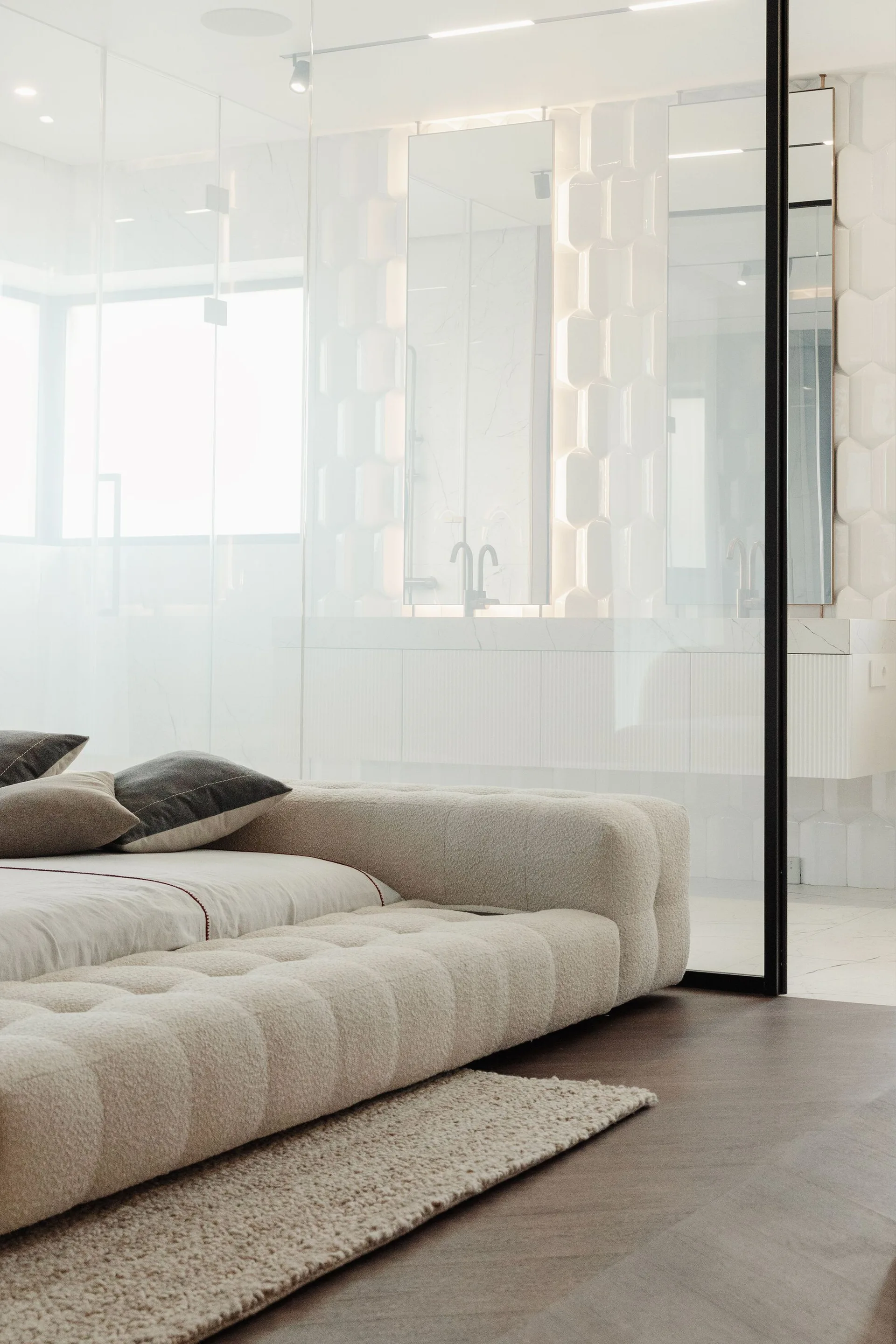 A modern bedroom features a low tufted platform bed on dark wood floors next to a black-framed glass partition revealing an ensuite bathroom.