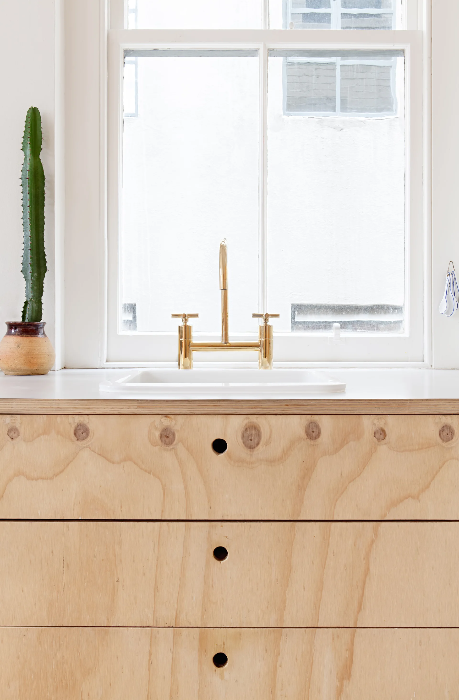 Plywood kitchen drawers with circular cut-out handles sit below a white countertop, a white undermount sink, and a brass bridge faucet in front of a window.