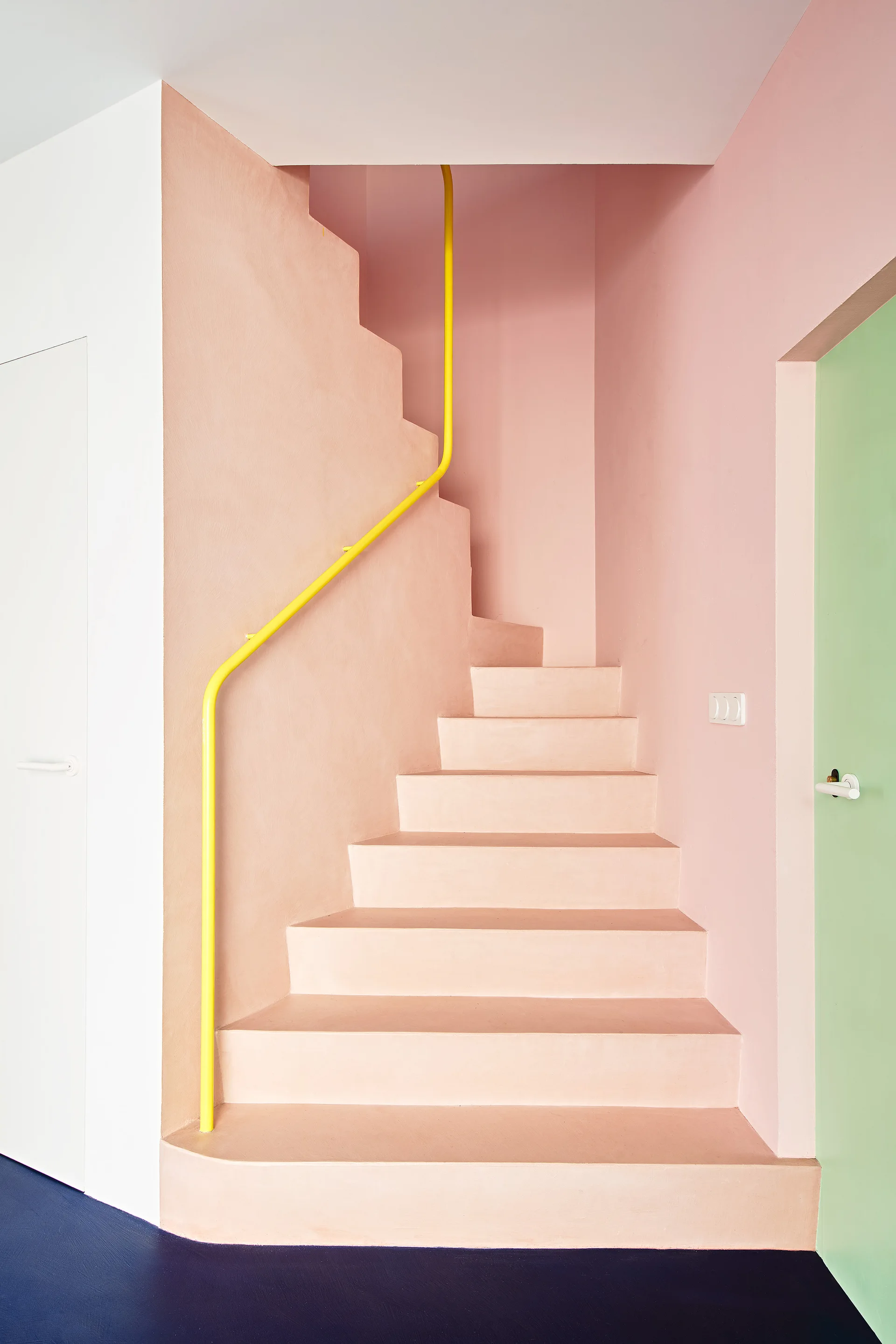 A minimalist staircase painted in soft peach features a bright yellow handrail, a navy floor, and a pastel green door on the right.