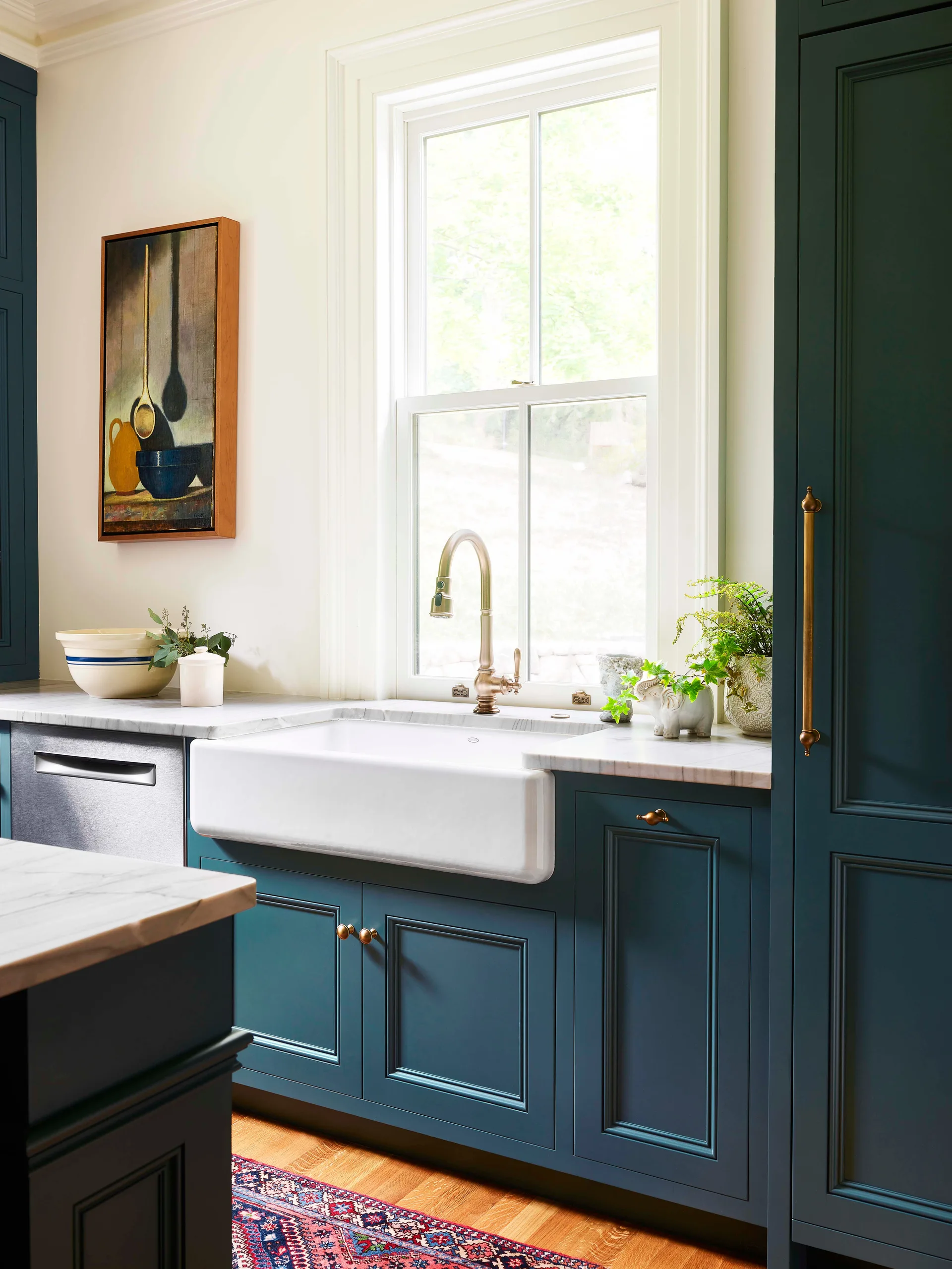 Deep green shaker cabinets surround a white ceramic farmhouse sink with a brass faucet beneath a window in a kitchen with marble countertops.