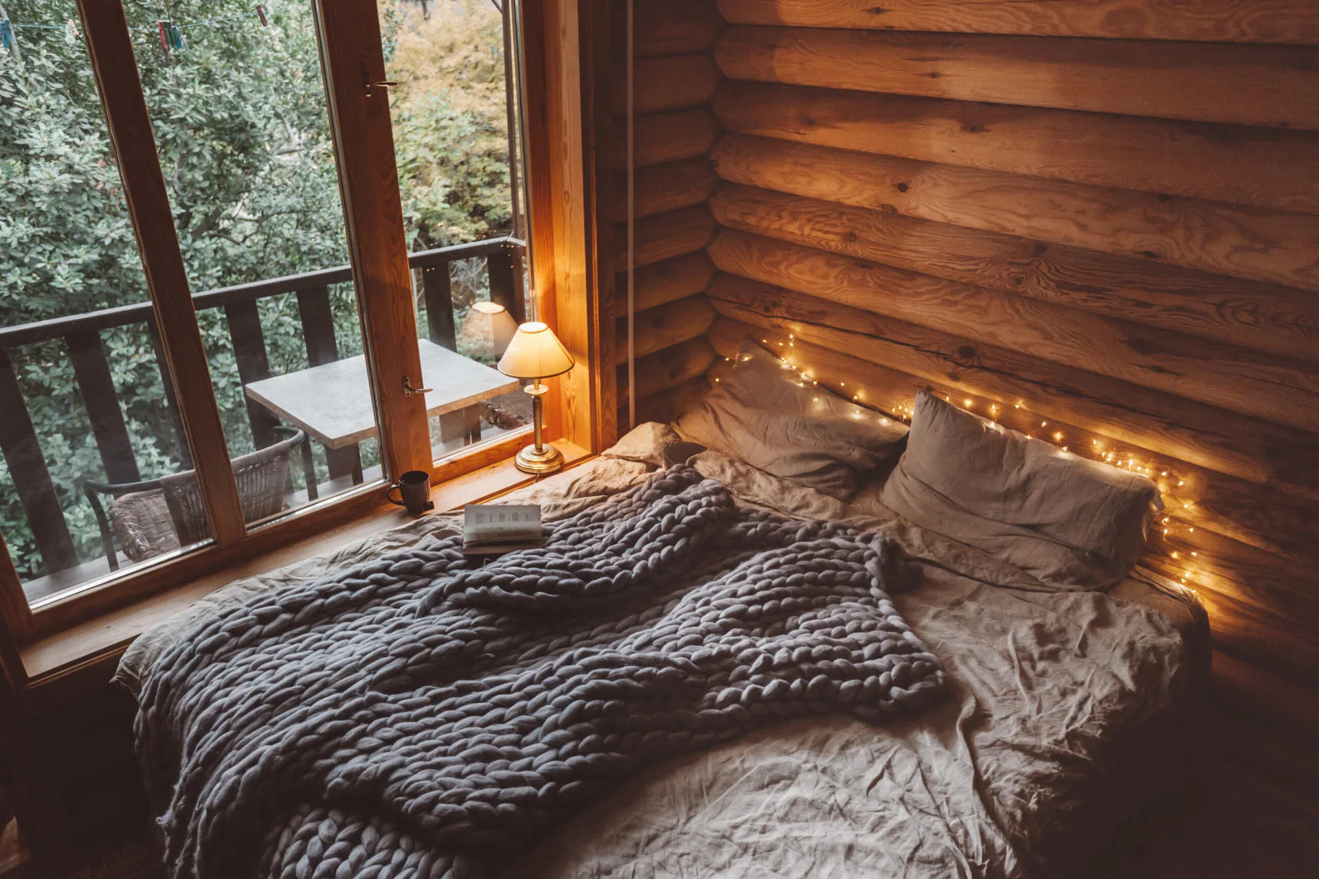 A bed with a heavy grey chunky knit blanket sits next to a glass door in a rustic cabin bedroom with log walls and warm lighting.