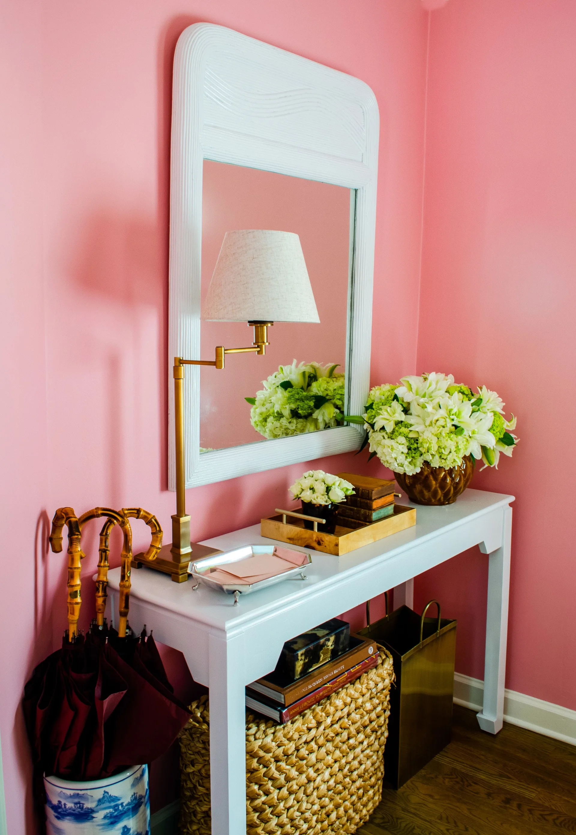 A white console table features a brass lamp, fresh flowers, and a white framed mirror set against a bright pink wall in an entryway.