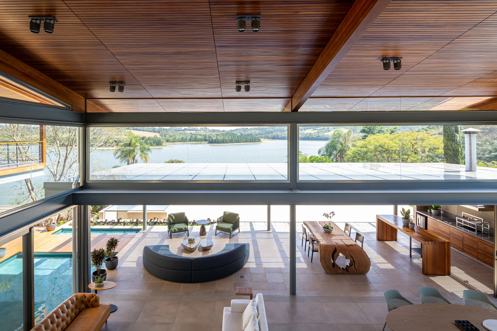 An open-concept living space with a wood-slat ceiling, curved grey sofa, and live-edge dining table overlooking a lake through large windows.