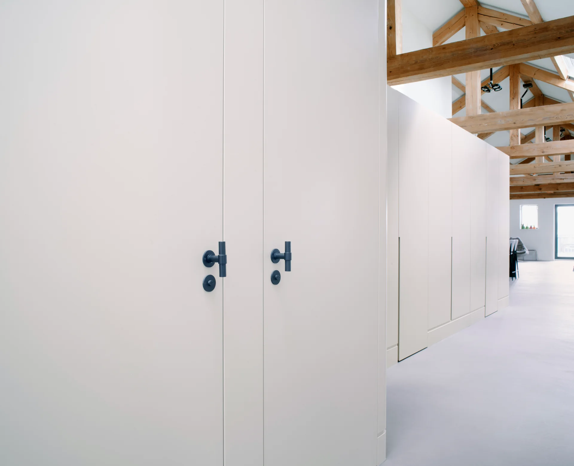 Matte white built-in storage cabinets with dark t-bar handles stand beneath an exposed wooden truss ceiling.