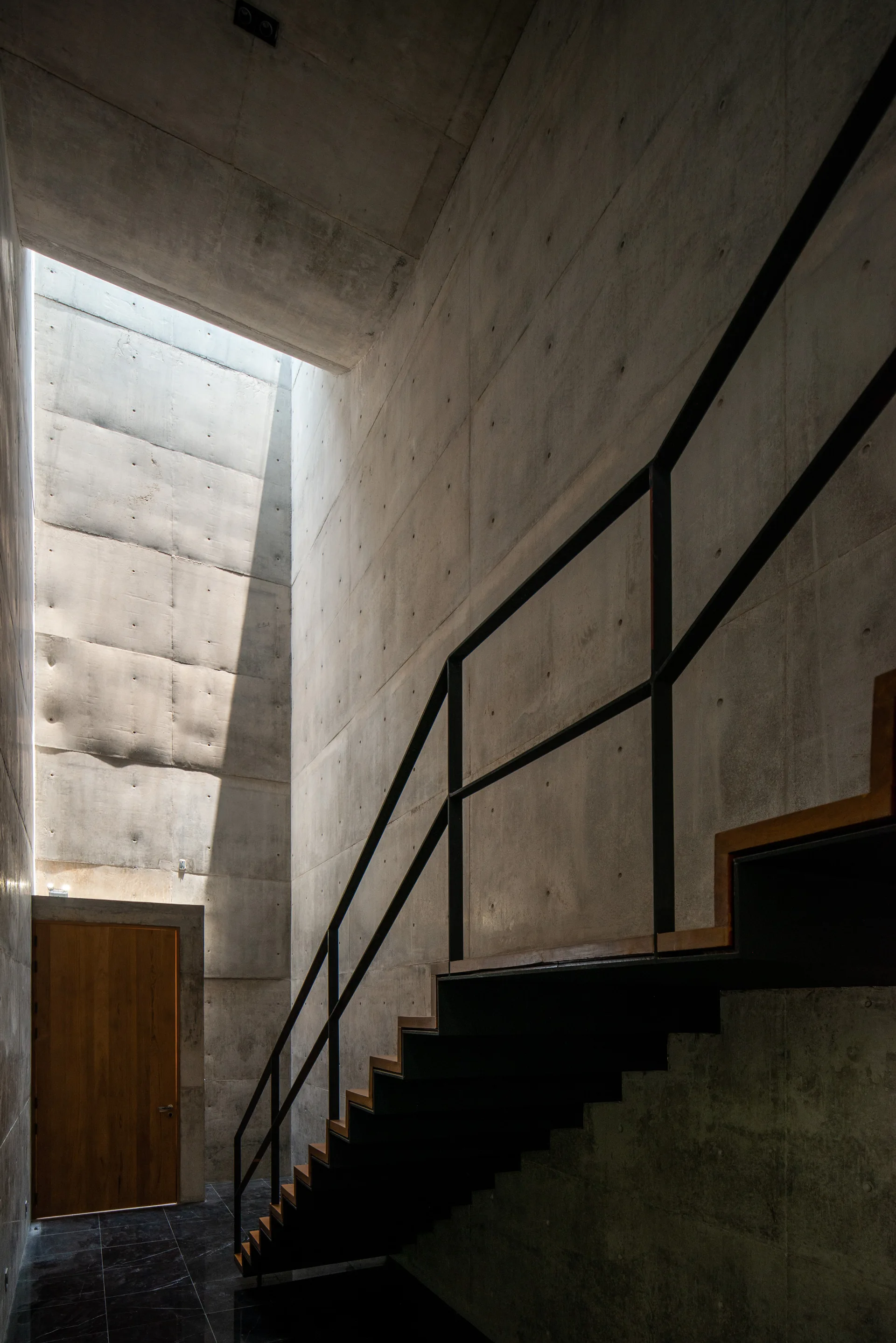 A minimalist concrete stairwell with a floating wooden staircase, black metal railing, a wood door at the bottom, and daylight pouring from a skylight.