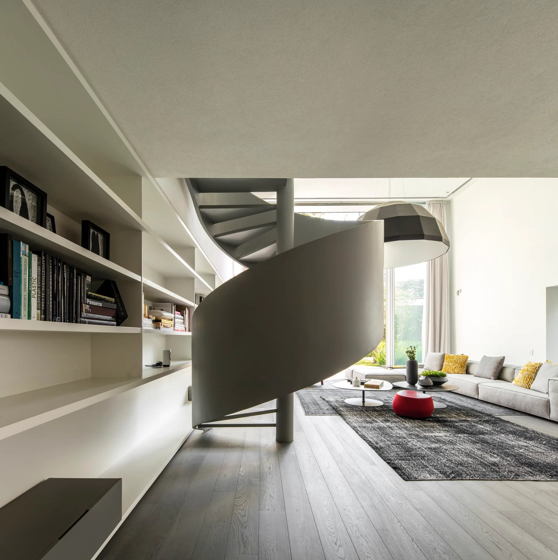 A contemporary living room contains a central dark gray spiral staircase, a large gray sectional sofa, and built-in white shelving on the left.