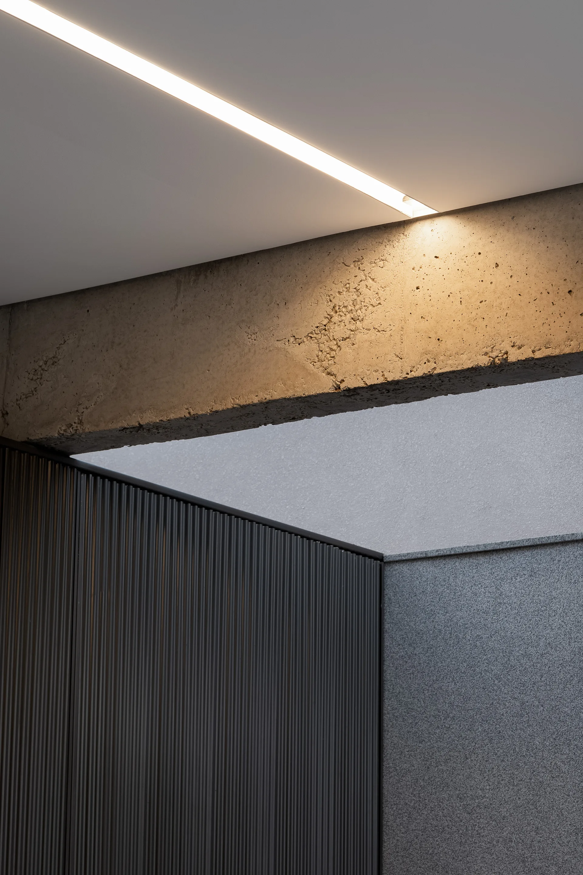 An architectural detail showing an exposed concrete beam beneath a white ceiling with a recessed linear light above vertical fluted wall paneling.