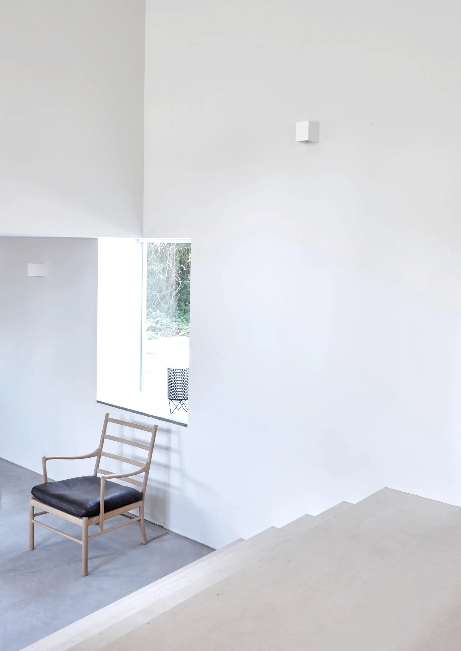 A minimalist white stairwell featuring a wooden armchair with a dark seat, concrete floors, and a small recessed square window.