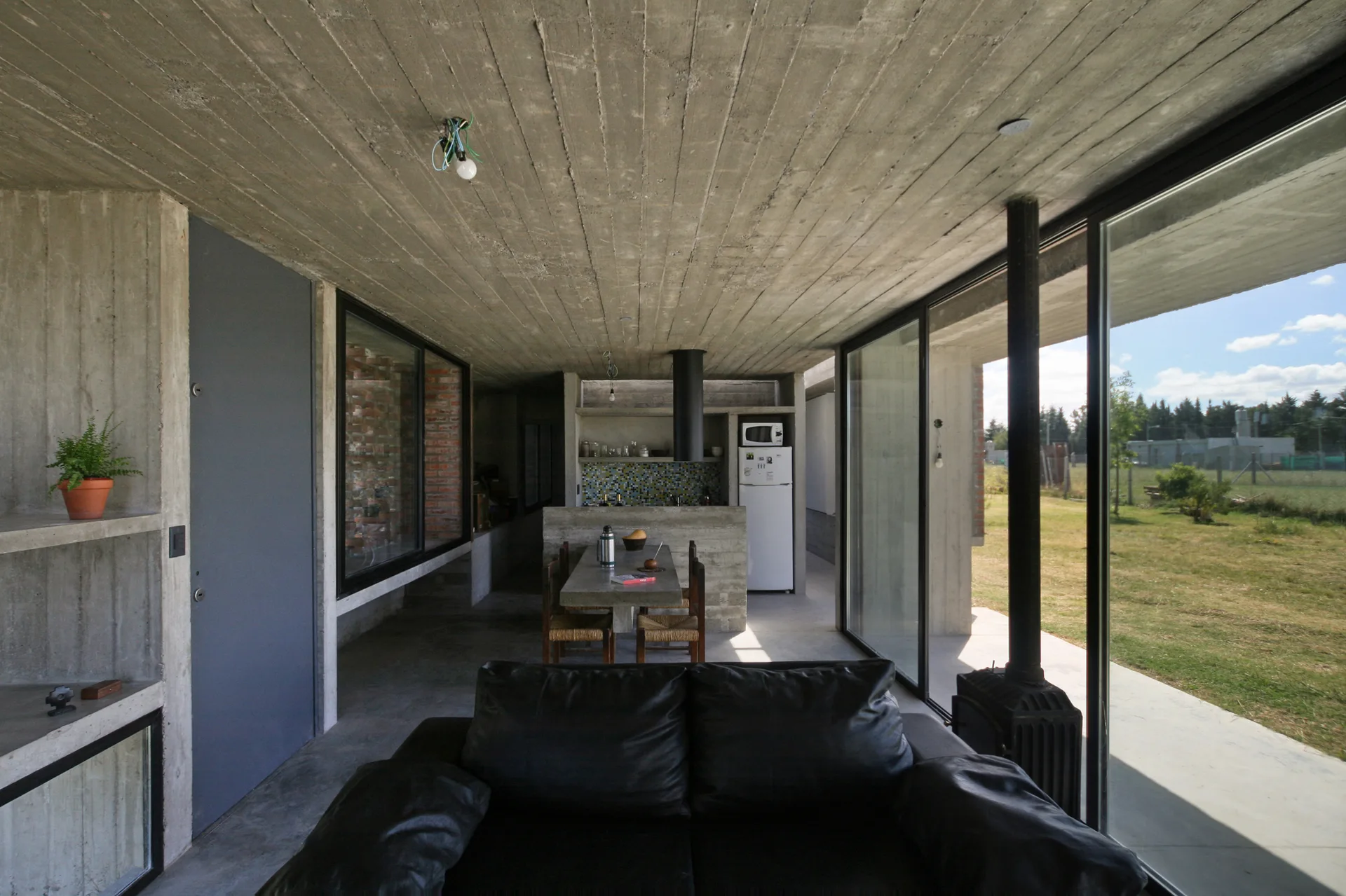 A minimalist open-concept living area features a black leather sofa, board-formed concrete walls, and large sliding glass doors opening to a grassy field.
