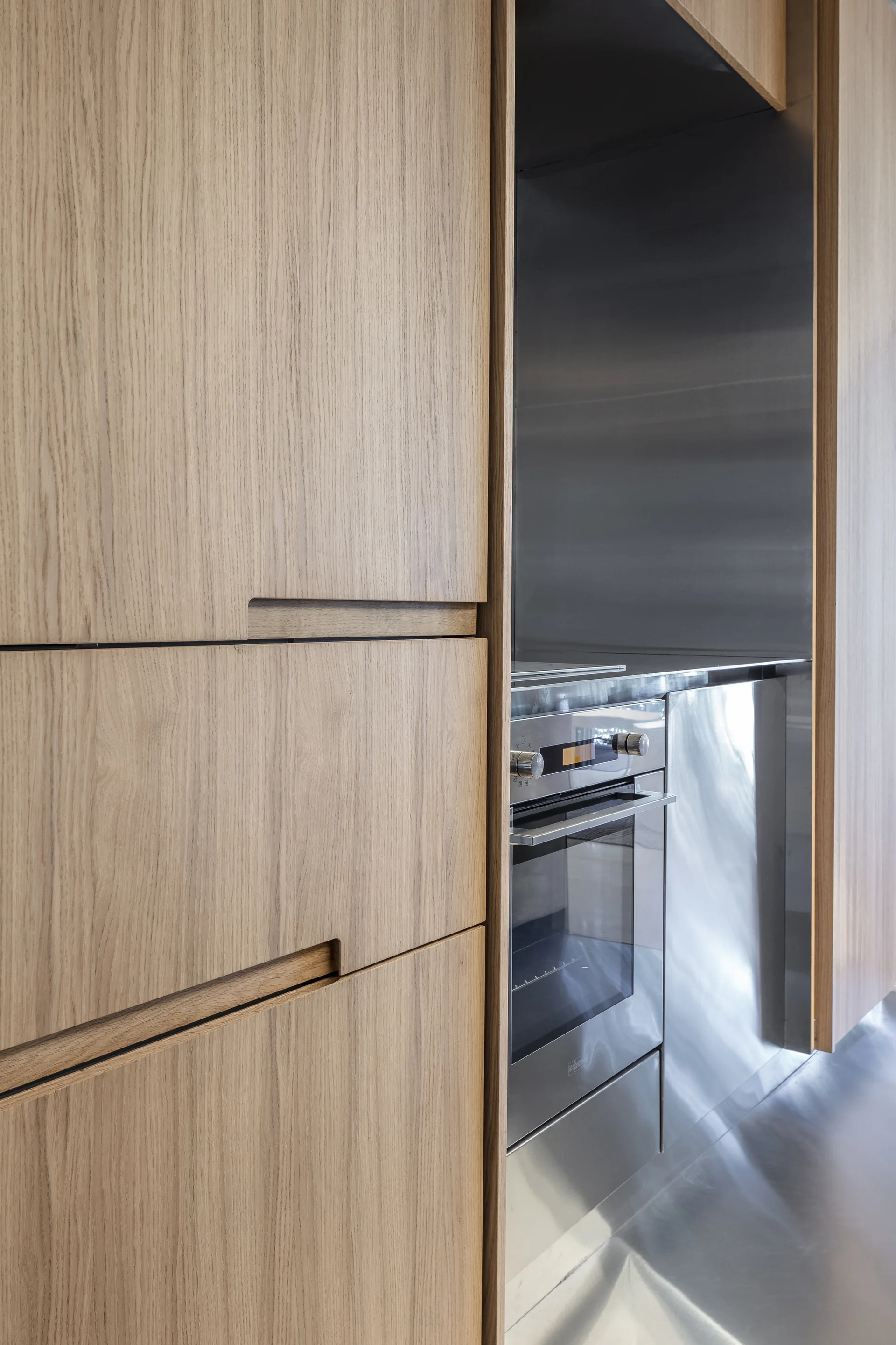 Close-up of flat-panel light wood kitchen cabinets featuring recessed handles alongside a built-in stainless steel oven and metal backsplash.