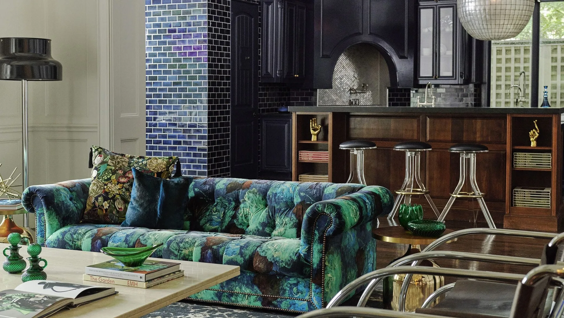 A moody open-plan room features a tufted green and blue floral velvet sofa near a glossy blue tiled column and a dark kitchen island.