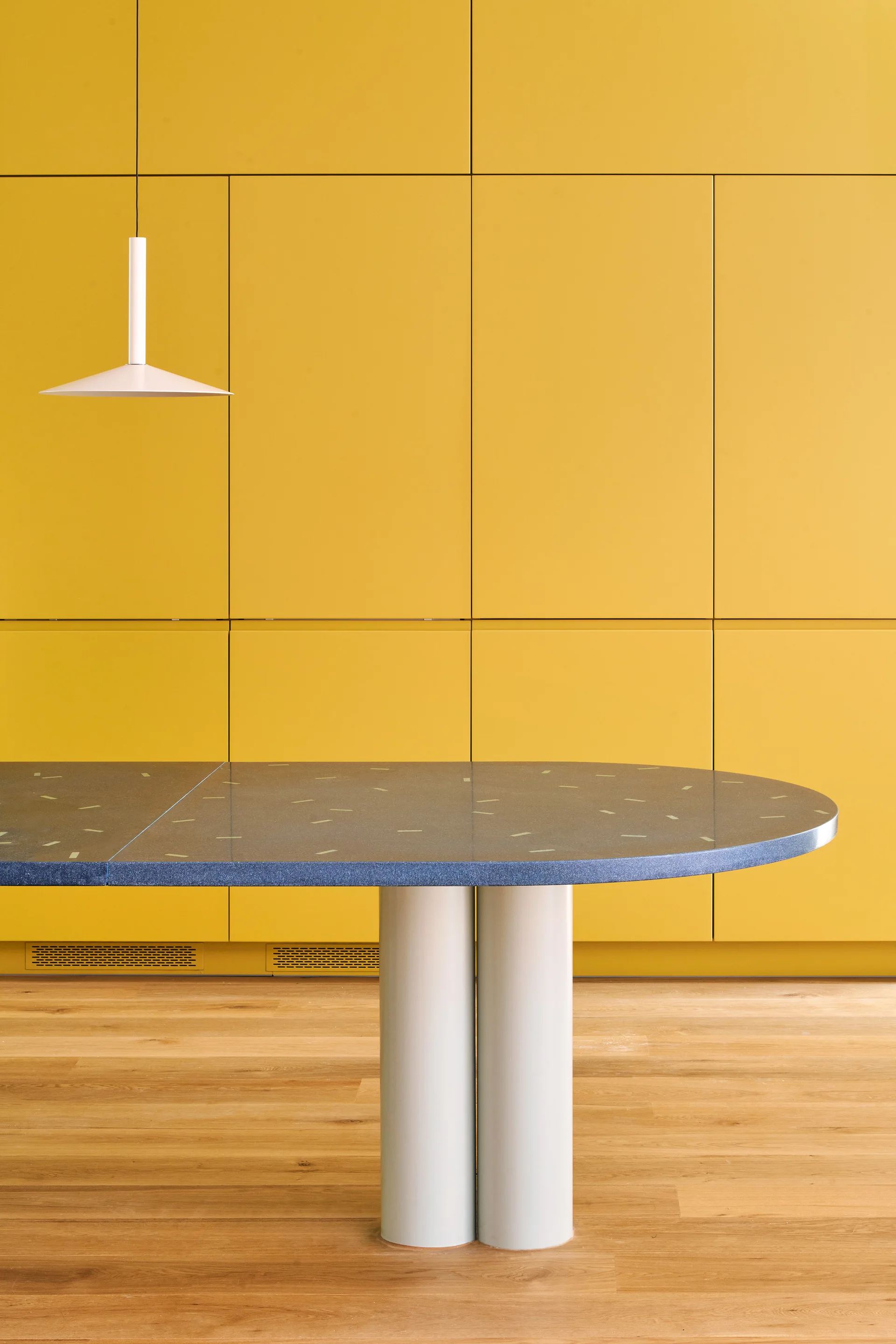 An elliptical blue speckled dining table on white column legs stands in front of a flat-front yellow cabinetry wall in a minimalist kitchen.