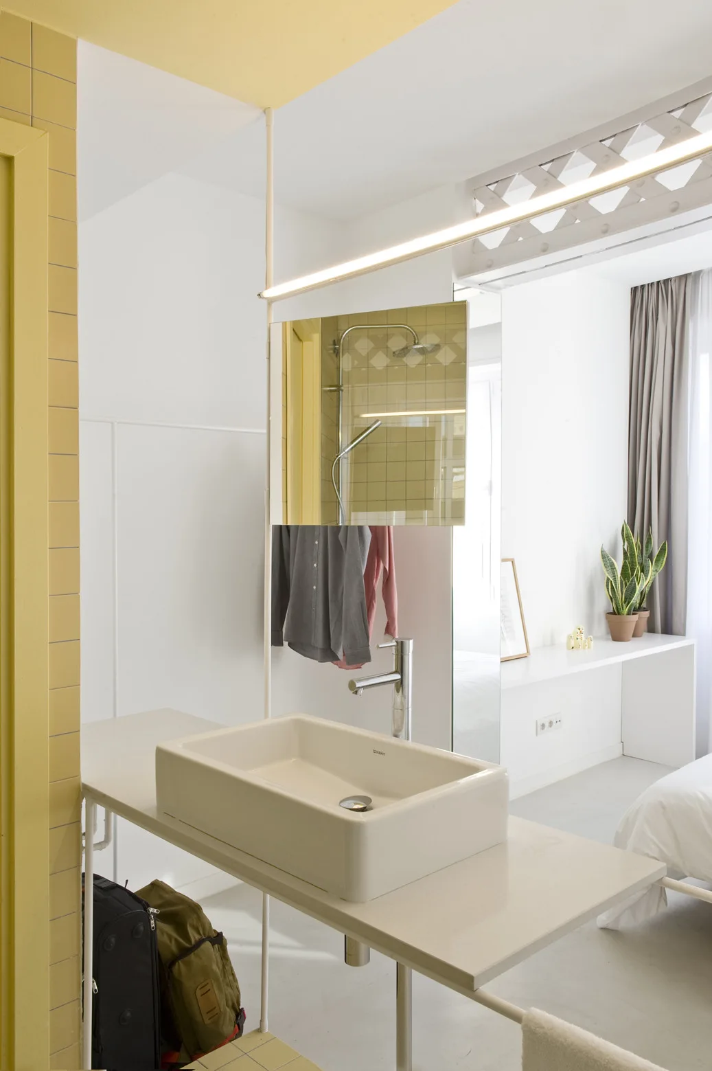 An open-concept bathroom vanity next to a bedroom features a rectangular vessel sink, a suspended mirror, and a yellow tiled wall.