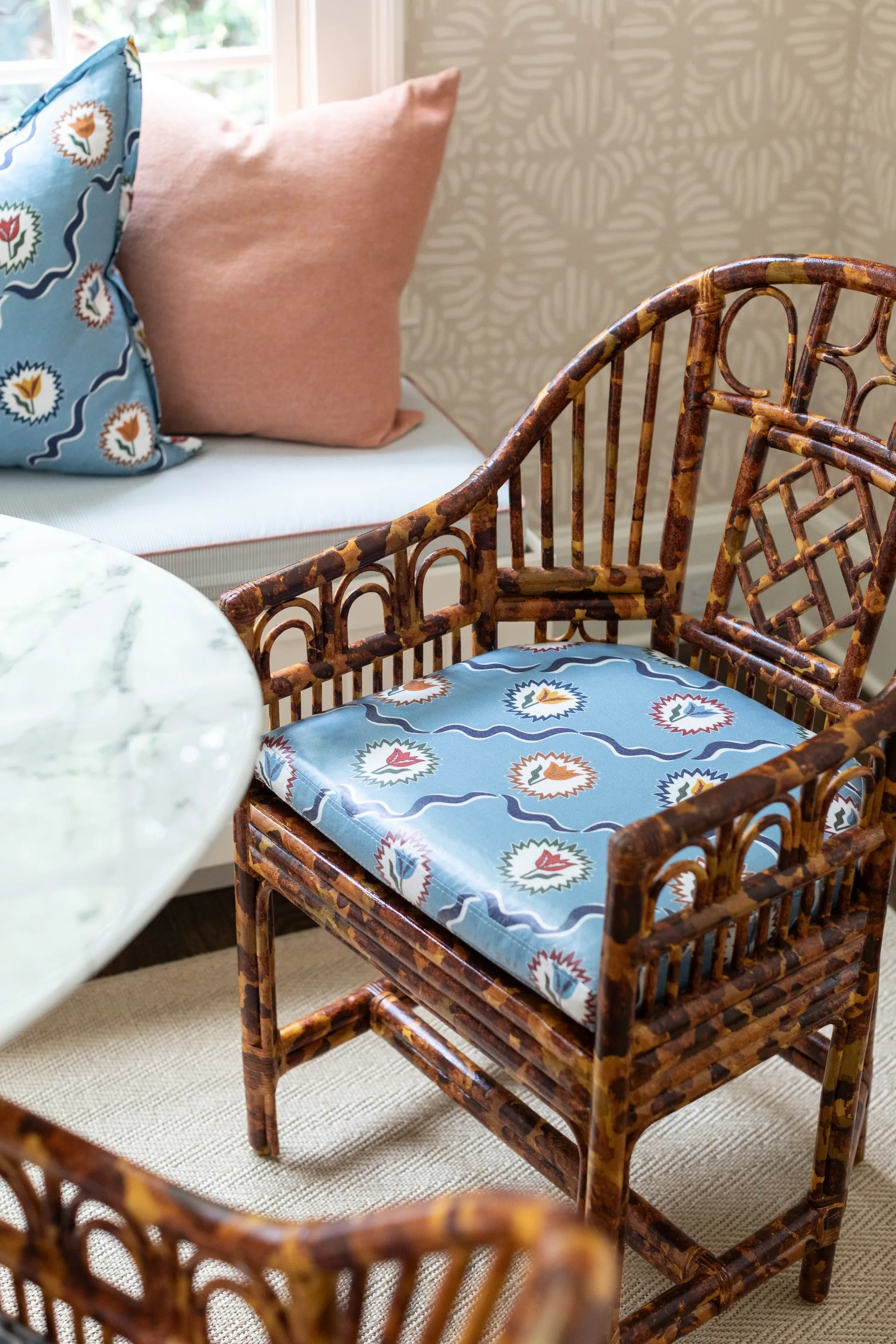 A mottled rattan armchair with a blue patterned cushion sits by a white marble dining table and a cushioned window seat.