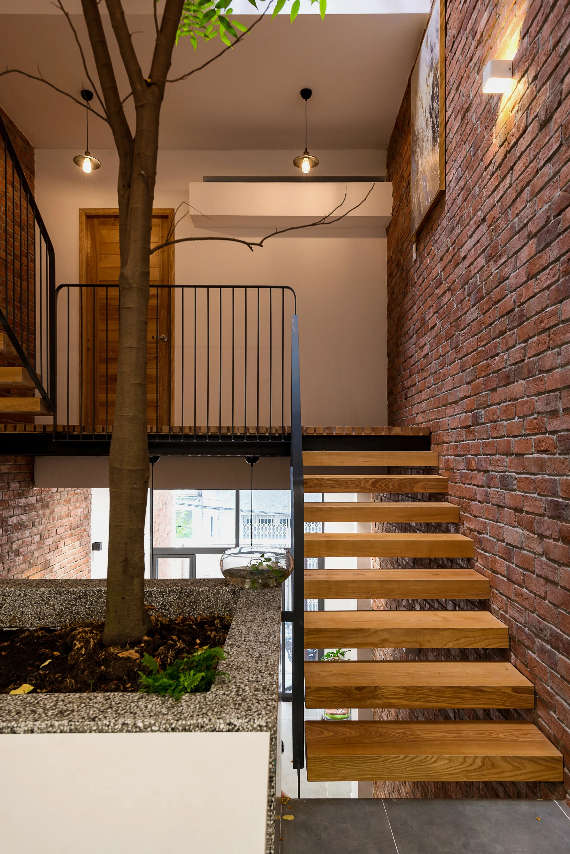 An industrial-style staircase with floating wood treads next to an exposed red brick wall and a tall indoor tree.