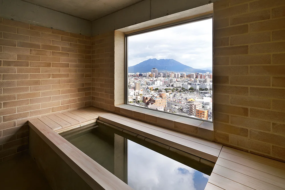 A minimalist bathroom featuring a wooden soaking tub, light brick walls, and a large window looking out onto a city and a distant mountain.