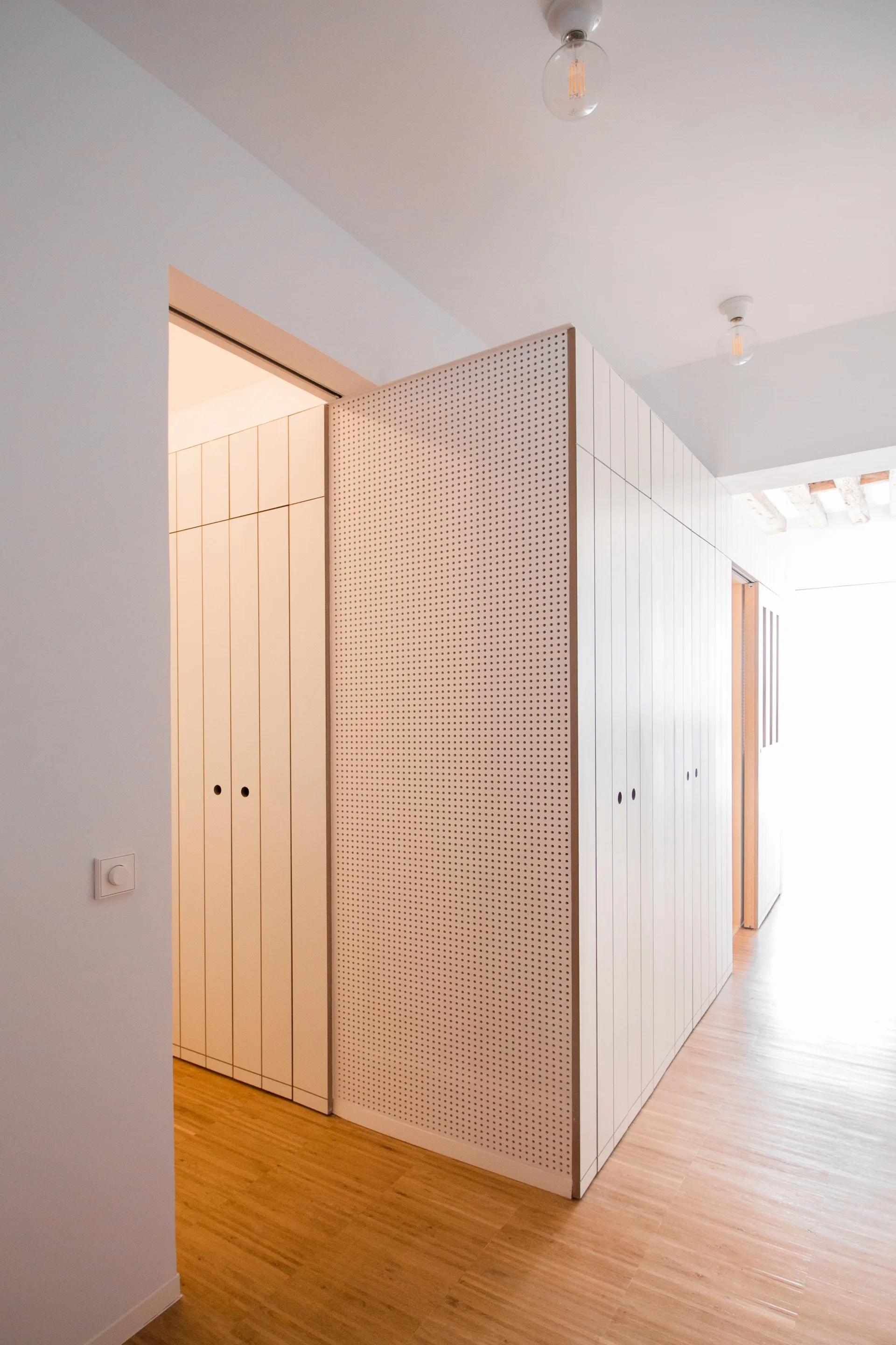 A minimalist white built-in cabinet unit with vertical grooves and a pegboard side panel stands in a hallway with light wood floors.