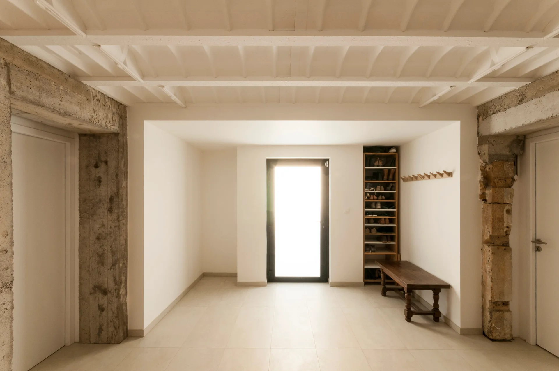 A minimalist entryway features white walls, painted ceiling beams, exposed stone pillars, a wooden bench, and a tall shoe rack near a bright glass door.