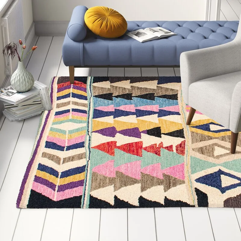 A multi-colored geometric patterned rug anchors a living room seating area next to a blue tufted daybed and a grey armchair on white floorboards.