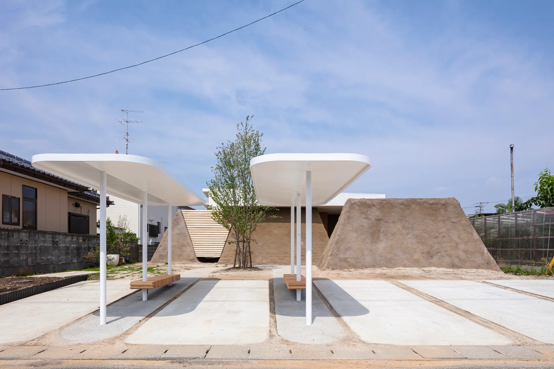 Two white minimalist canopies with wooden benches stand in a concrete courtyard framed by textured earth mounds and a central tree.
