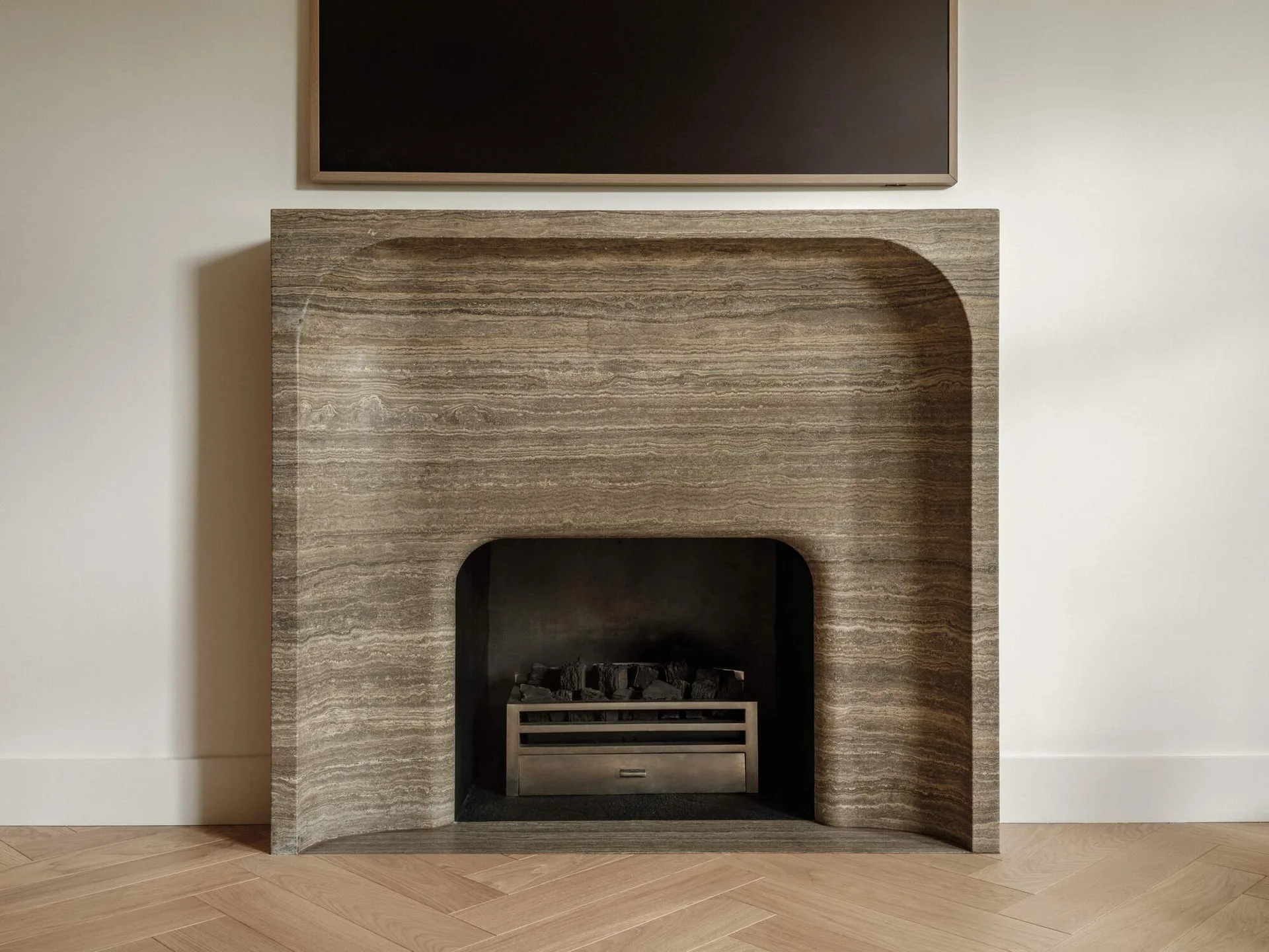 A minimalist stone fireplace with horizontal veining sits on a herringbone patterned wood floor beneath a wall-mounted television.