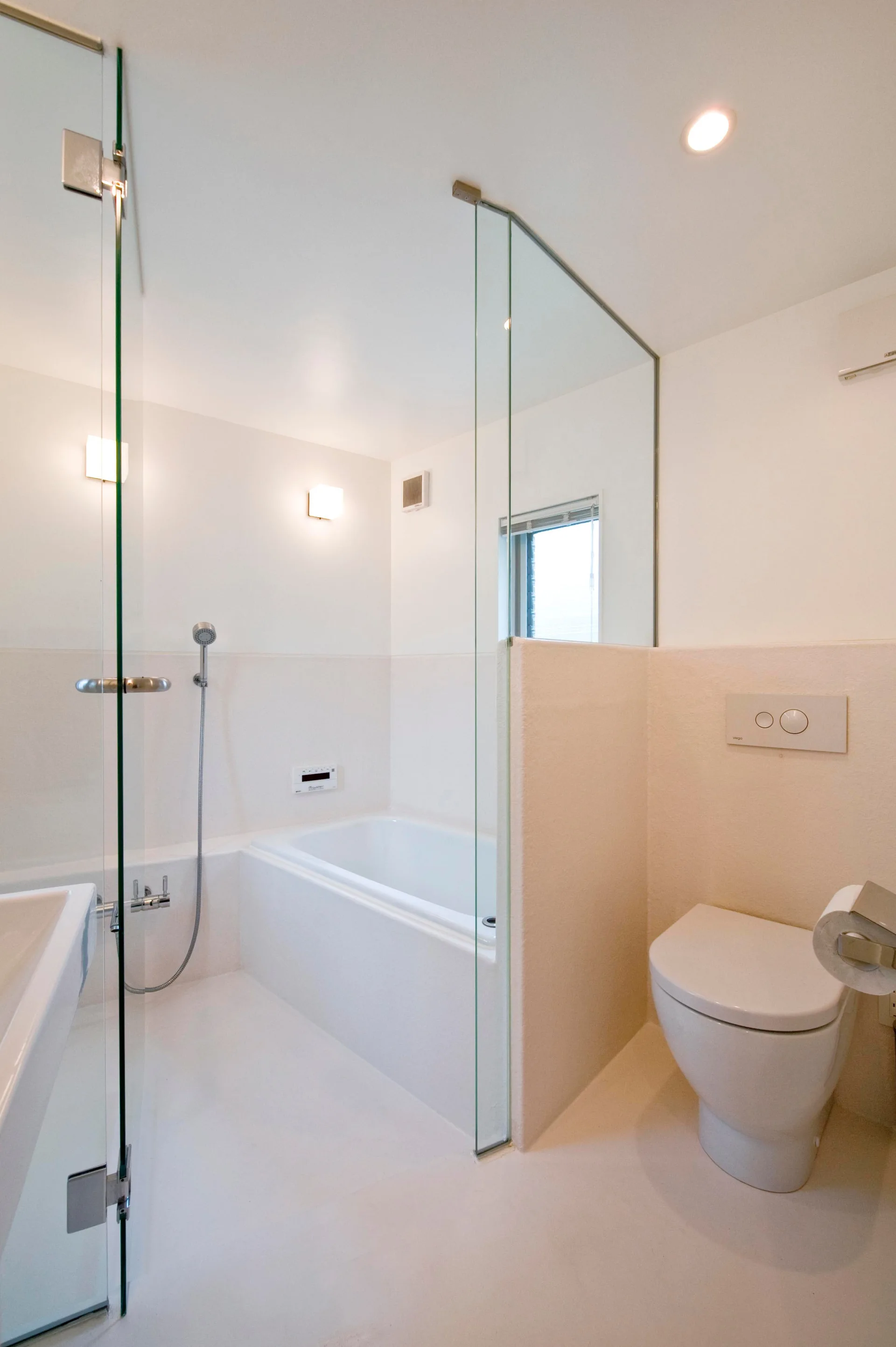 A minimalist white bathroom featuring a bathtub, a wall-mounted toilet, and a partition wall made of solid plaster and clear glass.