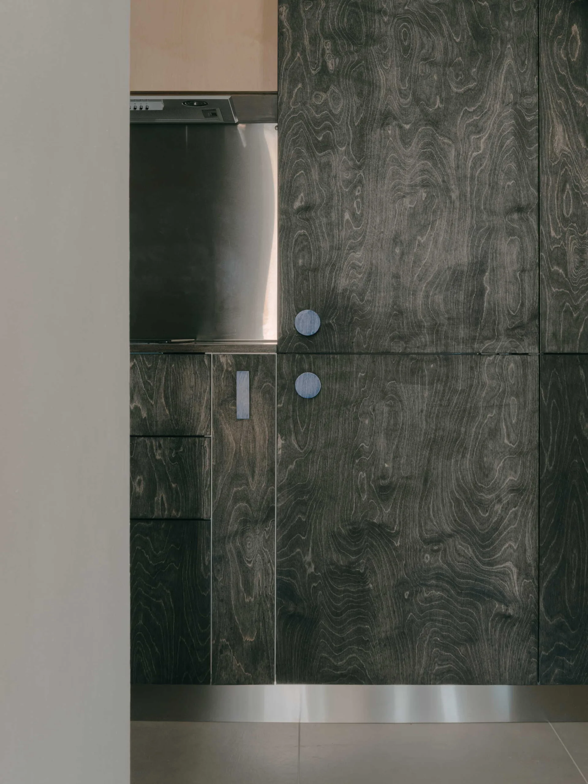 A close-up of dark charcoal stained wood kitchen cabinets with swirling grain patterns, circular blue pulls, and a stainless steel backsplash.