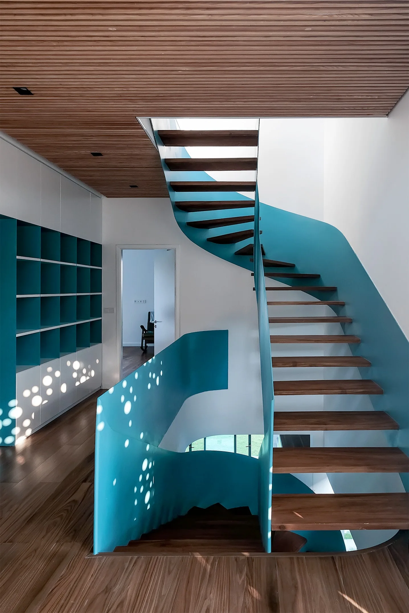 A modern blue metal spiral staircase with natural wood treads stands below a slatted timber ceiling.