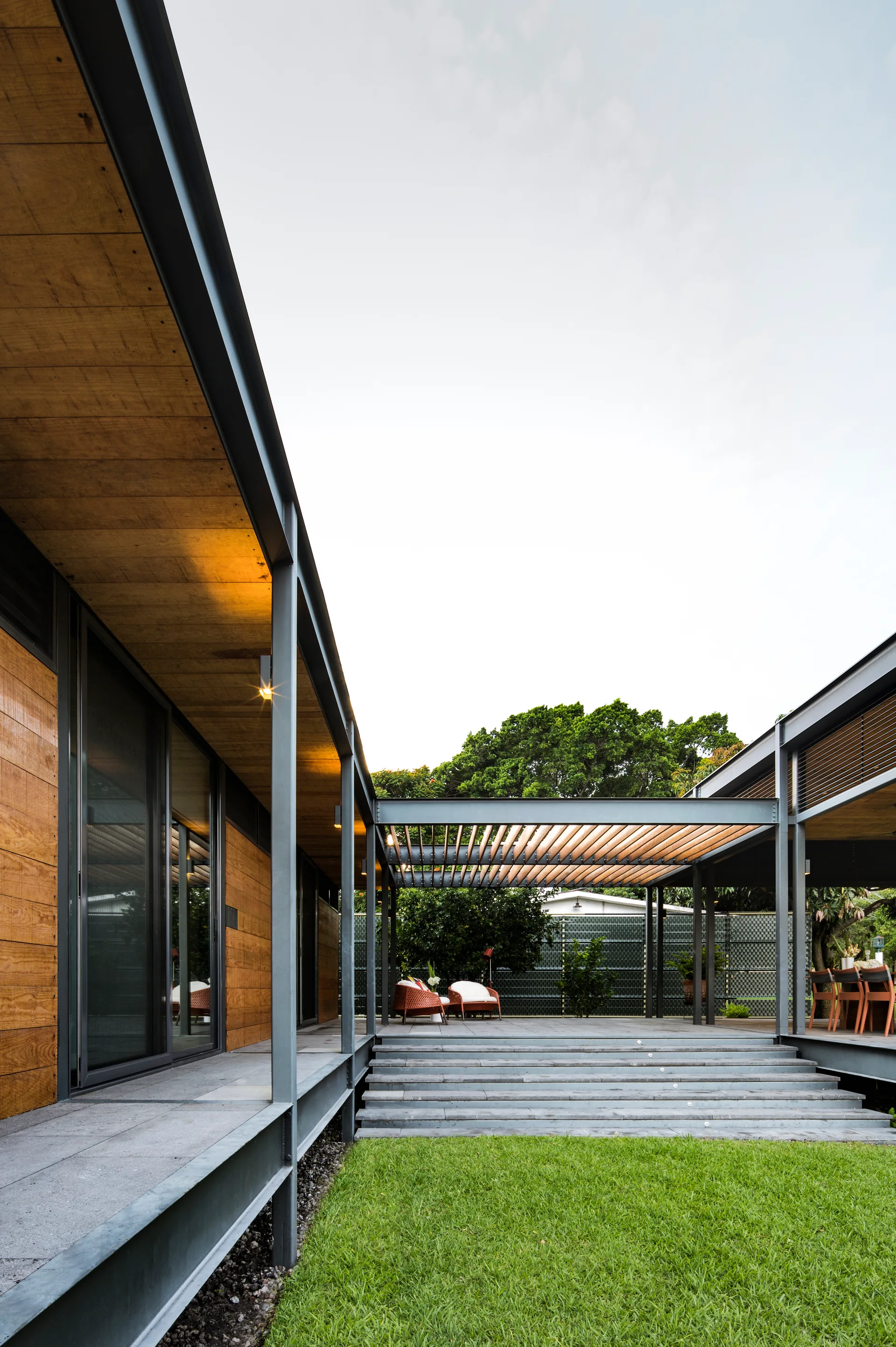 A modern home exterior features a steel-framed patio with a slatted wood pergola, grey stone steps, and a green grass lawn.