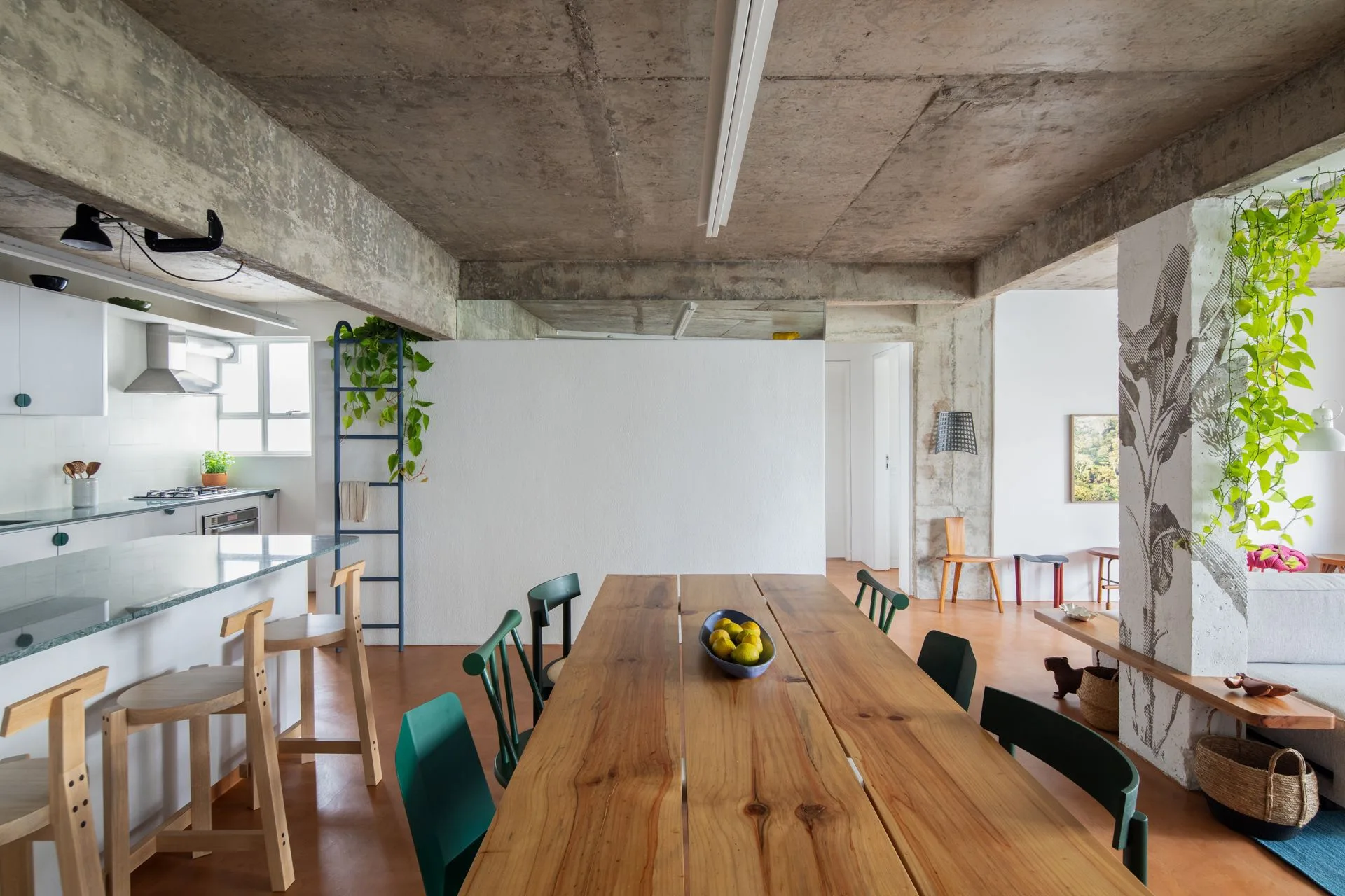 An open plan dining area features a long wooden table, green chairs, an exposed concrete ceiling, and a column painted with a leaf motif.