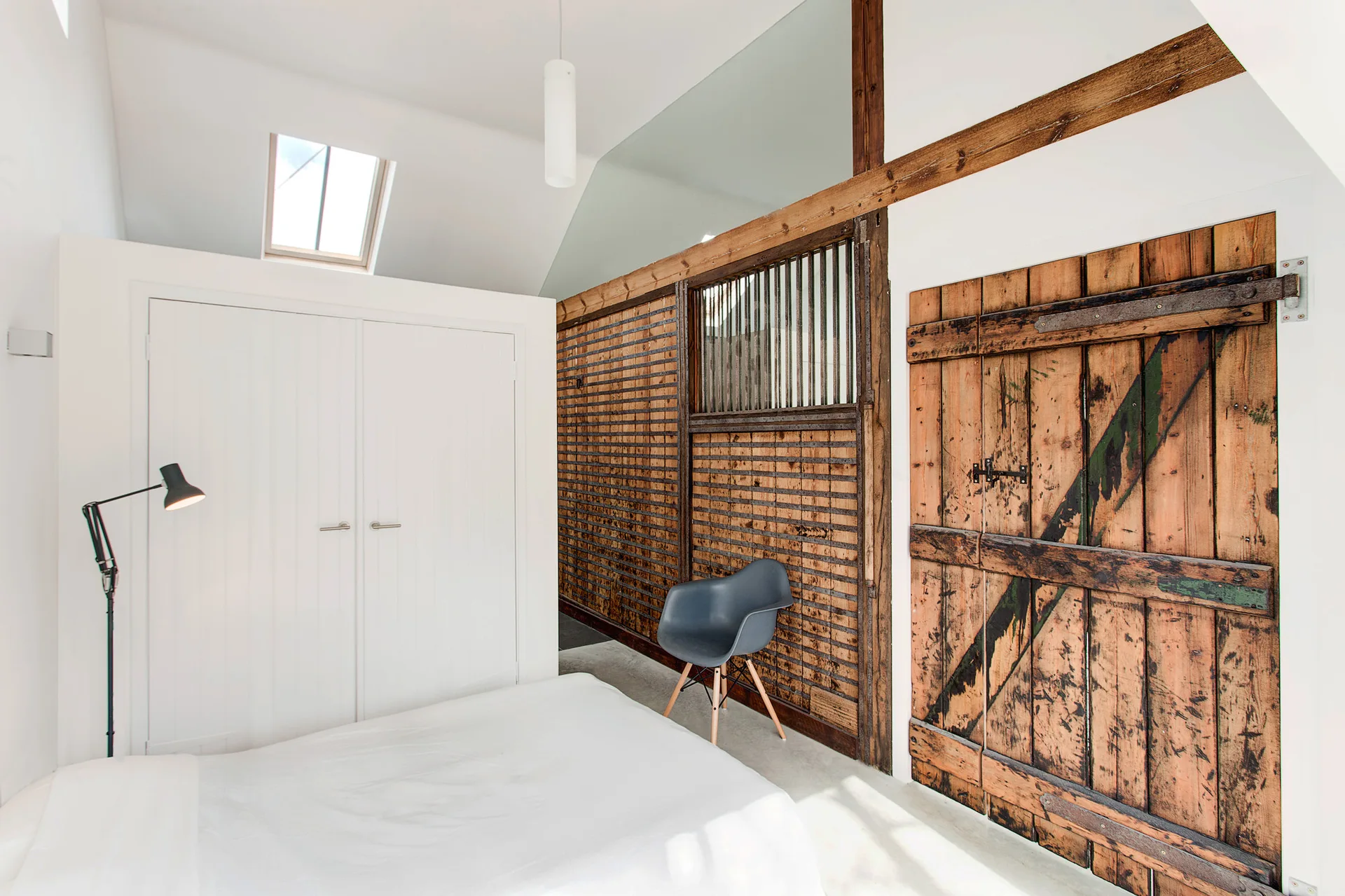 A minimalist bedroom features a white bed, a white built-in closet, a grey shell chair, and a rustic reclaimed barn wood accent wall under a sloped ceiling.