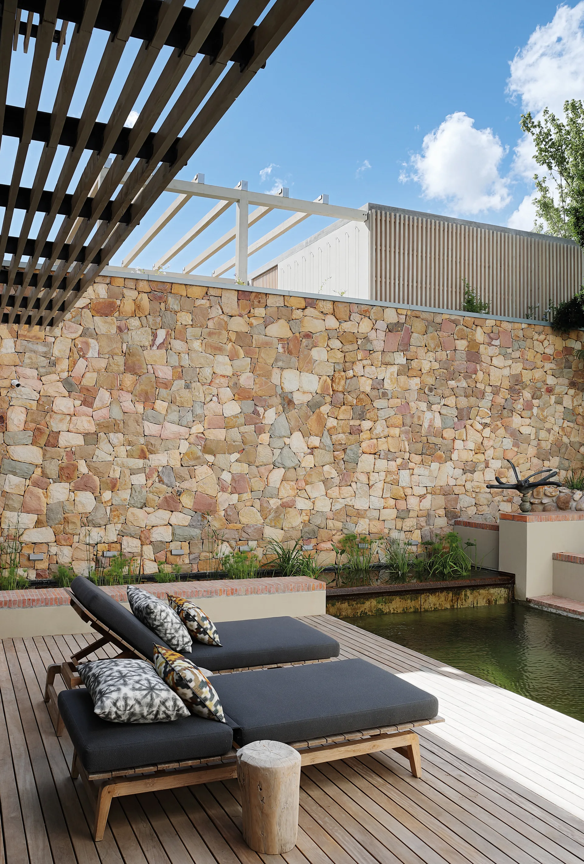 Two gray sun loungers sit on a wooden deck next to a garden pond and a tall stacked stone feature wall under a slatted pergola.