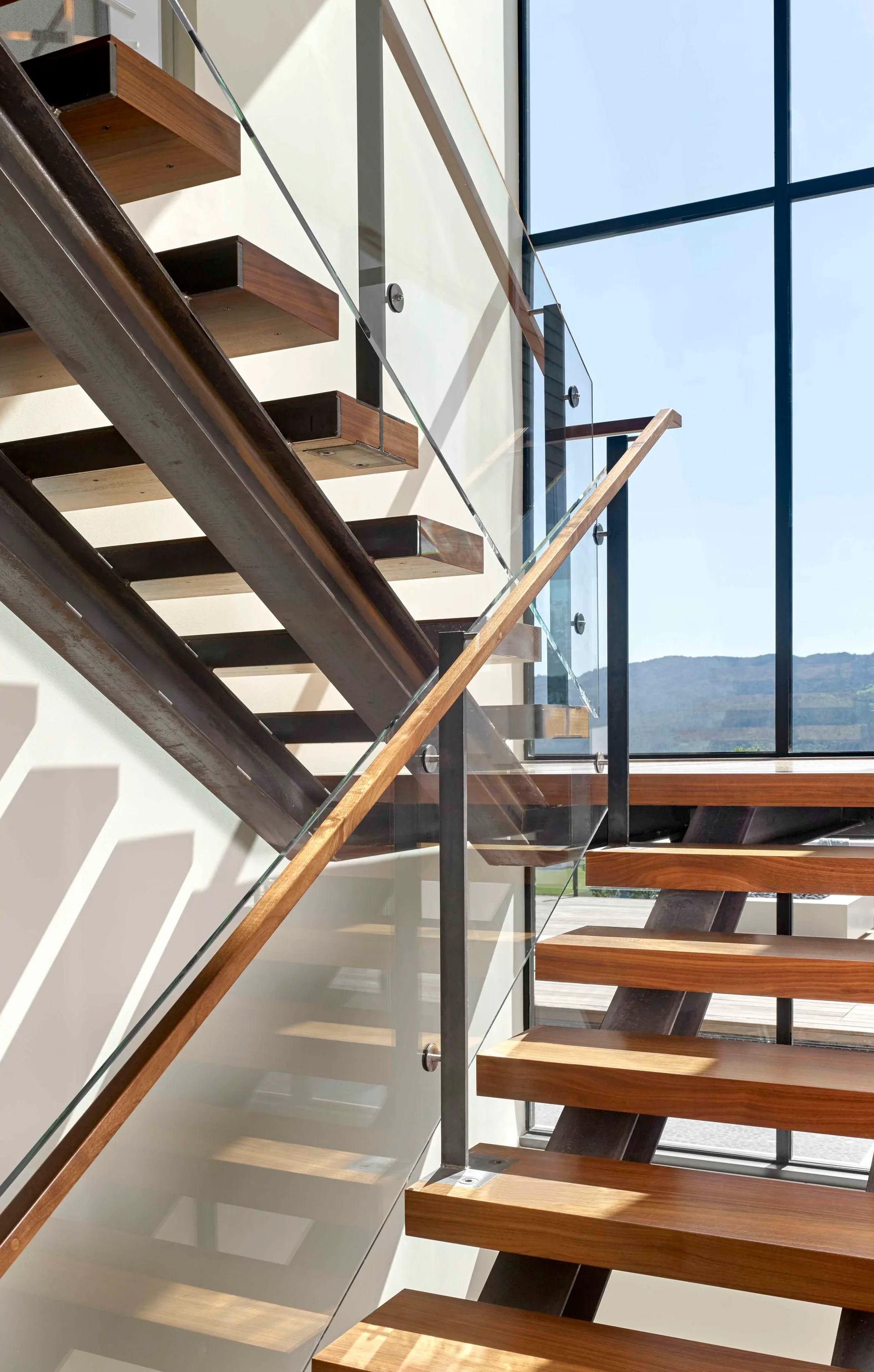 A modern staircase with thick floating wooden treads, a dark metal frame, and clear glass railings set against a large window with natural light.